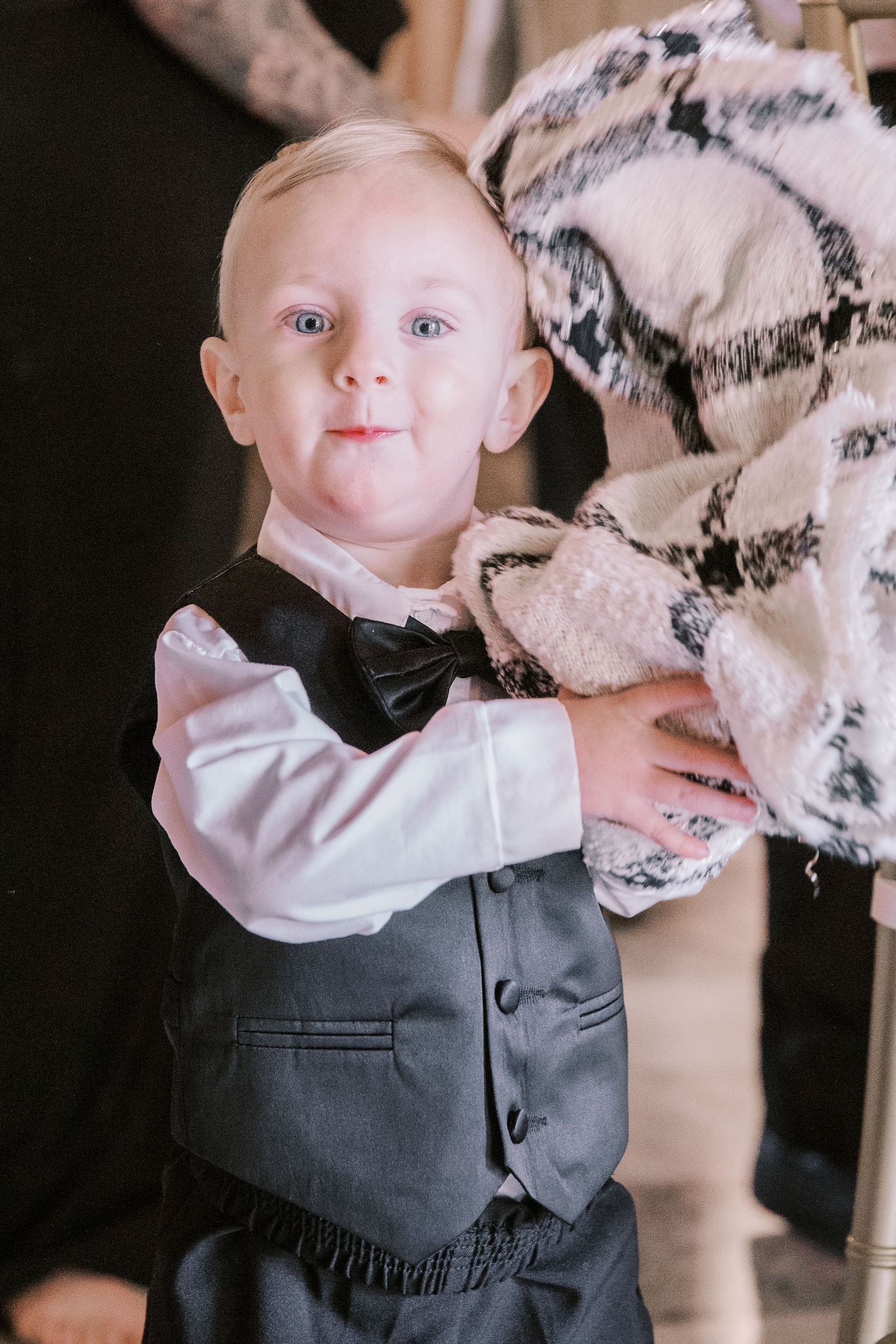 A young child dressed in formal attire with a black bow tie and grey vest holds a plush toy.