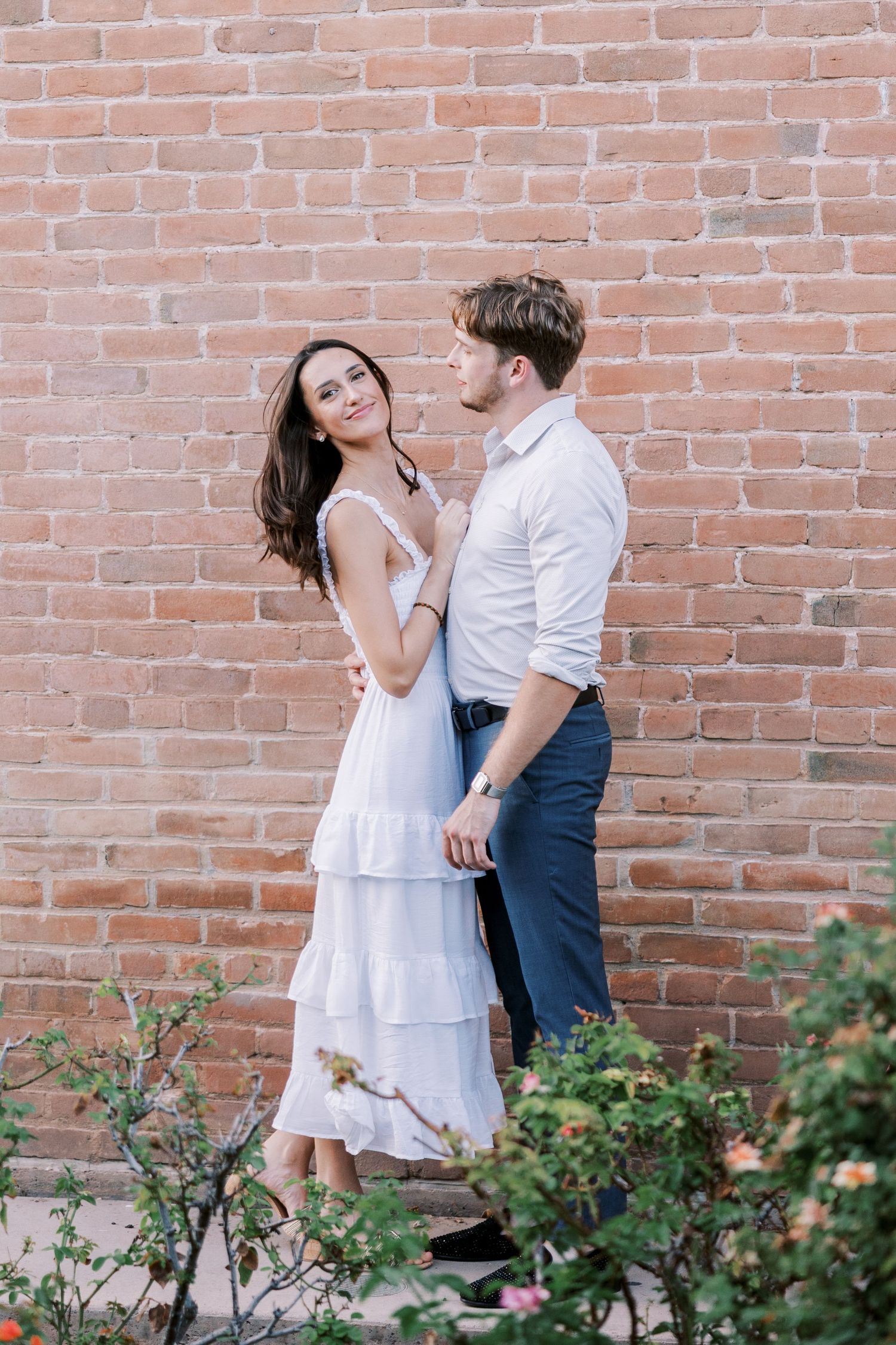 A couple in white clothing shares tender moments against a rustic brick wall with blooming roses.