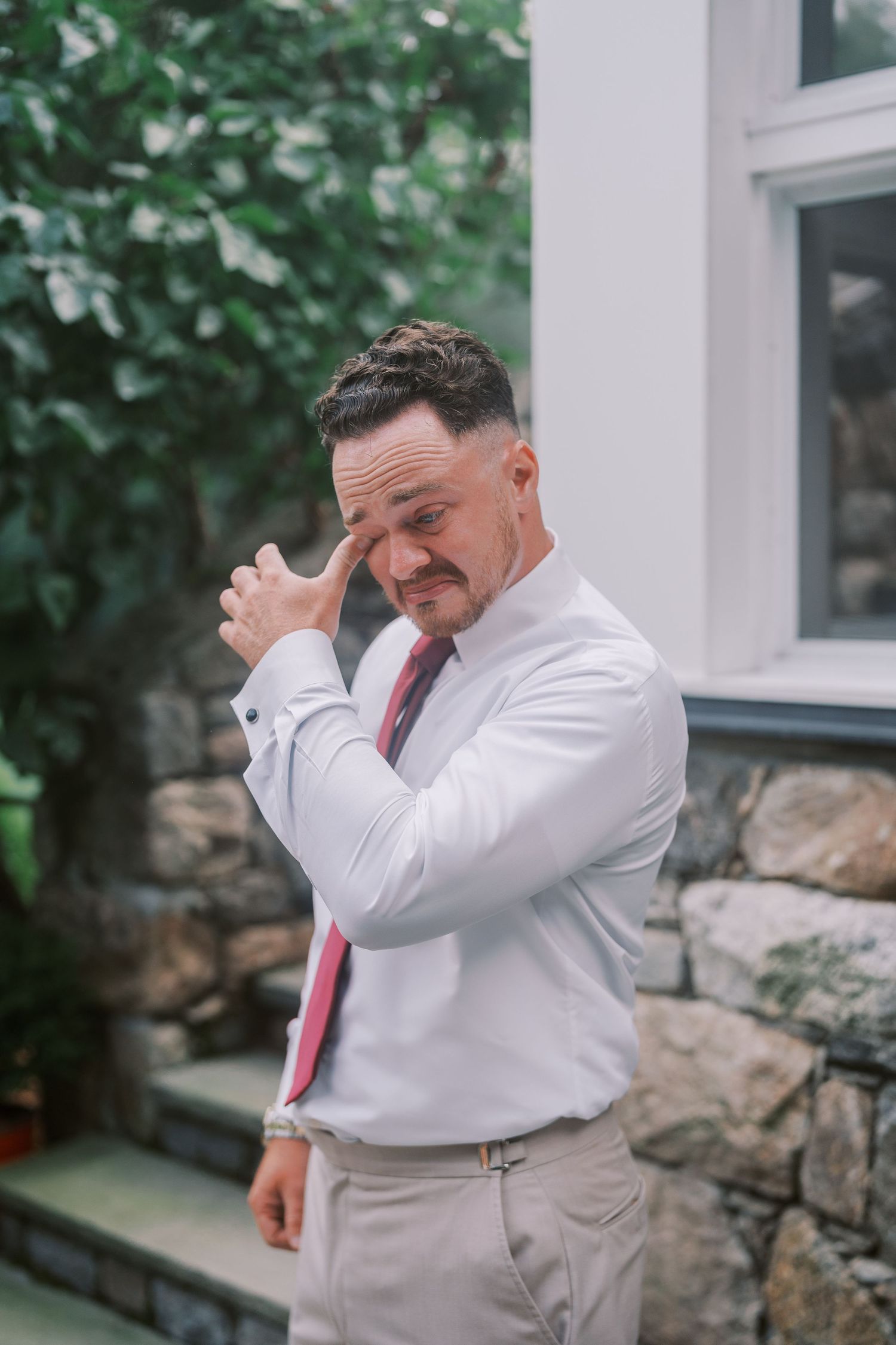 A person in a white dress shirt and red tie adjusts their sleeve while standing outdoors near greenery and stone wall.