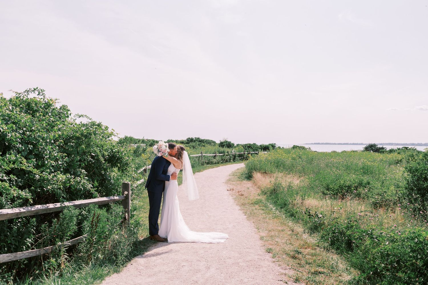 A romantic couple embraces on a scenic coastal path lined with green foliage and wooden railings.