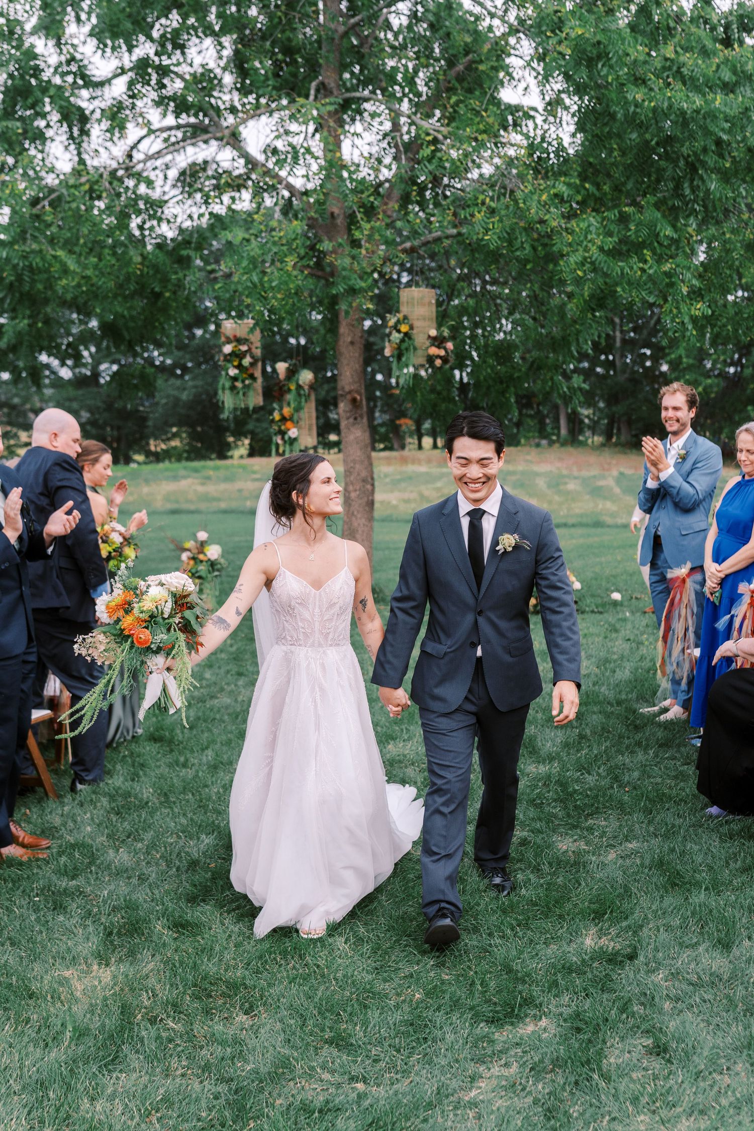 Newlyweds walk hand in hand on grass while wedding guests cheer.