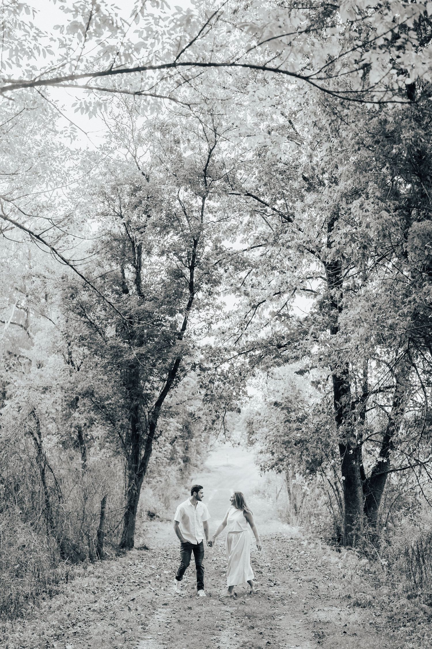 A black and white photo of a couple walking through a dreamy tree-covered path.