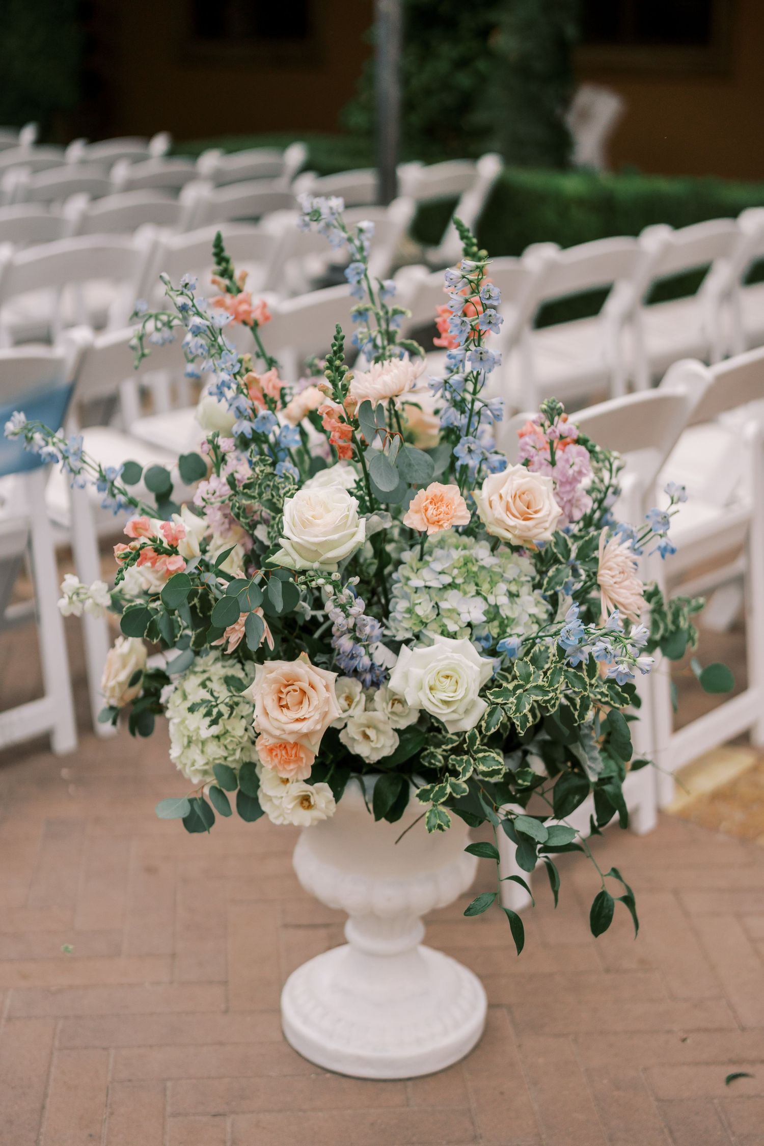 White and peach floral arrangements in classic urns line a wedding ceremony aisle on a brick patio.