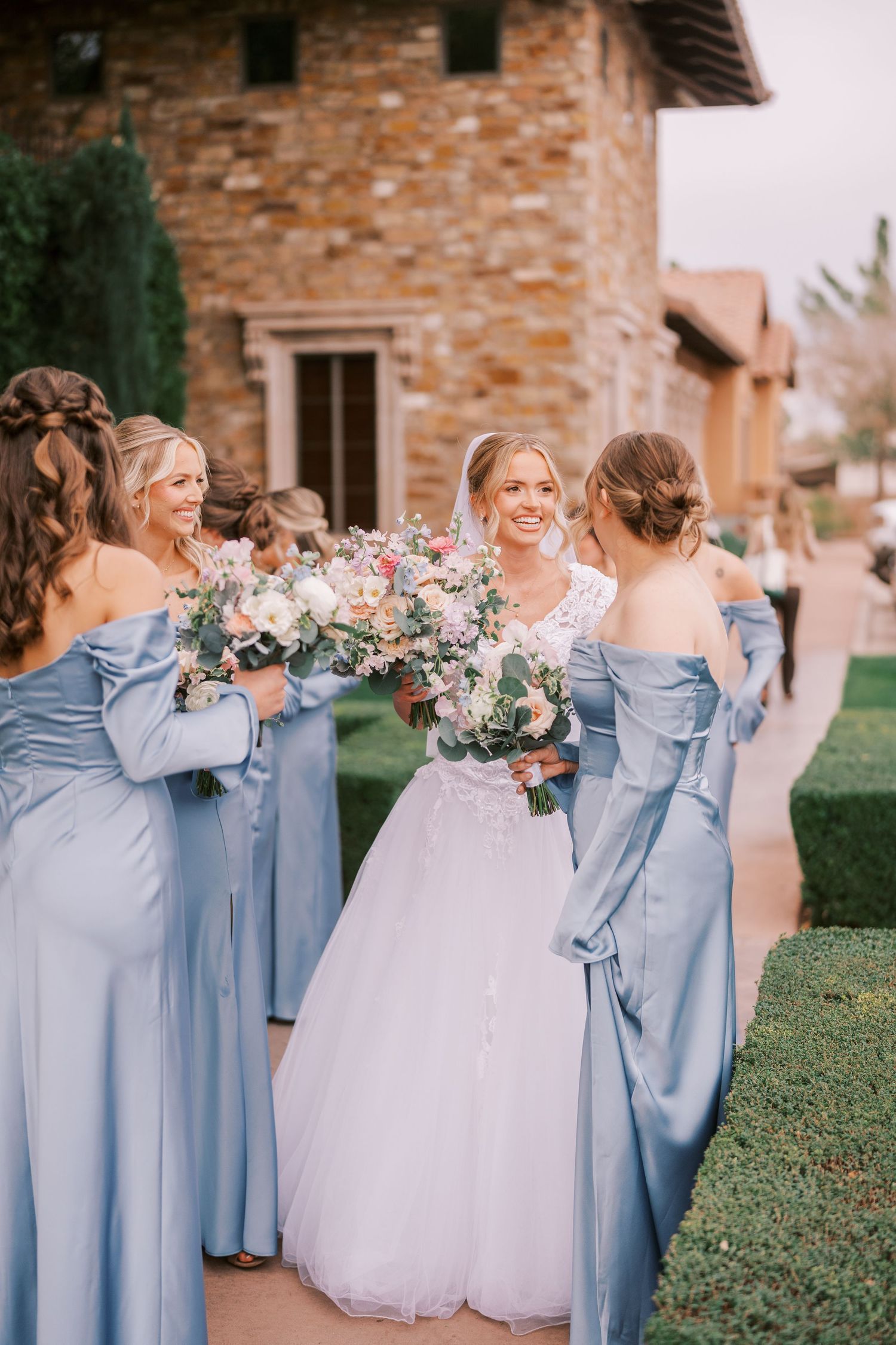 Wedding party in powder blue bridesmaid dresses gather around bride holding pink and white floral bouquets outside stone venue.