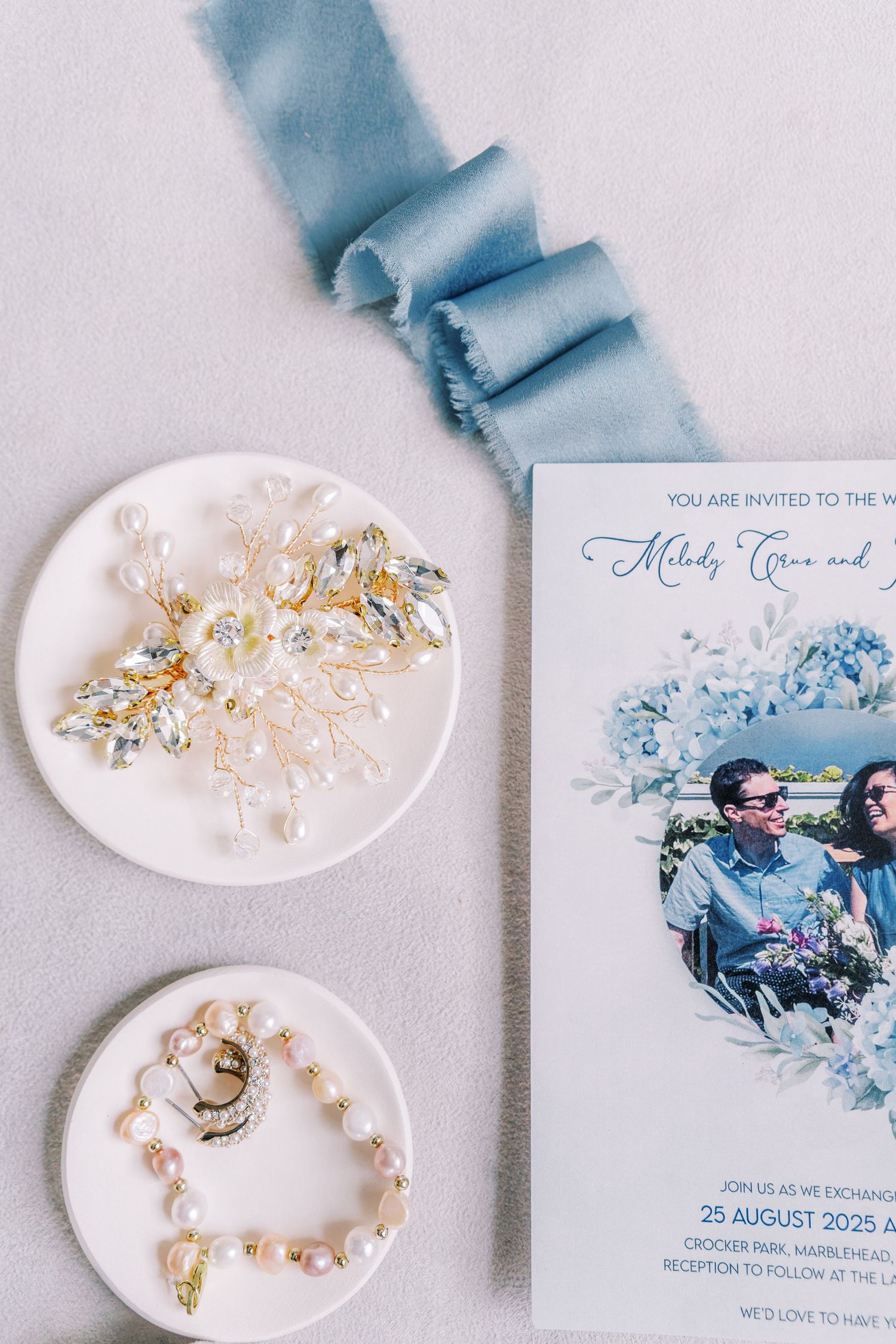 Wedding stationery in blue tones with pearl accessories and floral elements on white background.