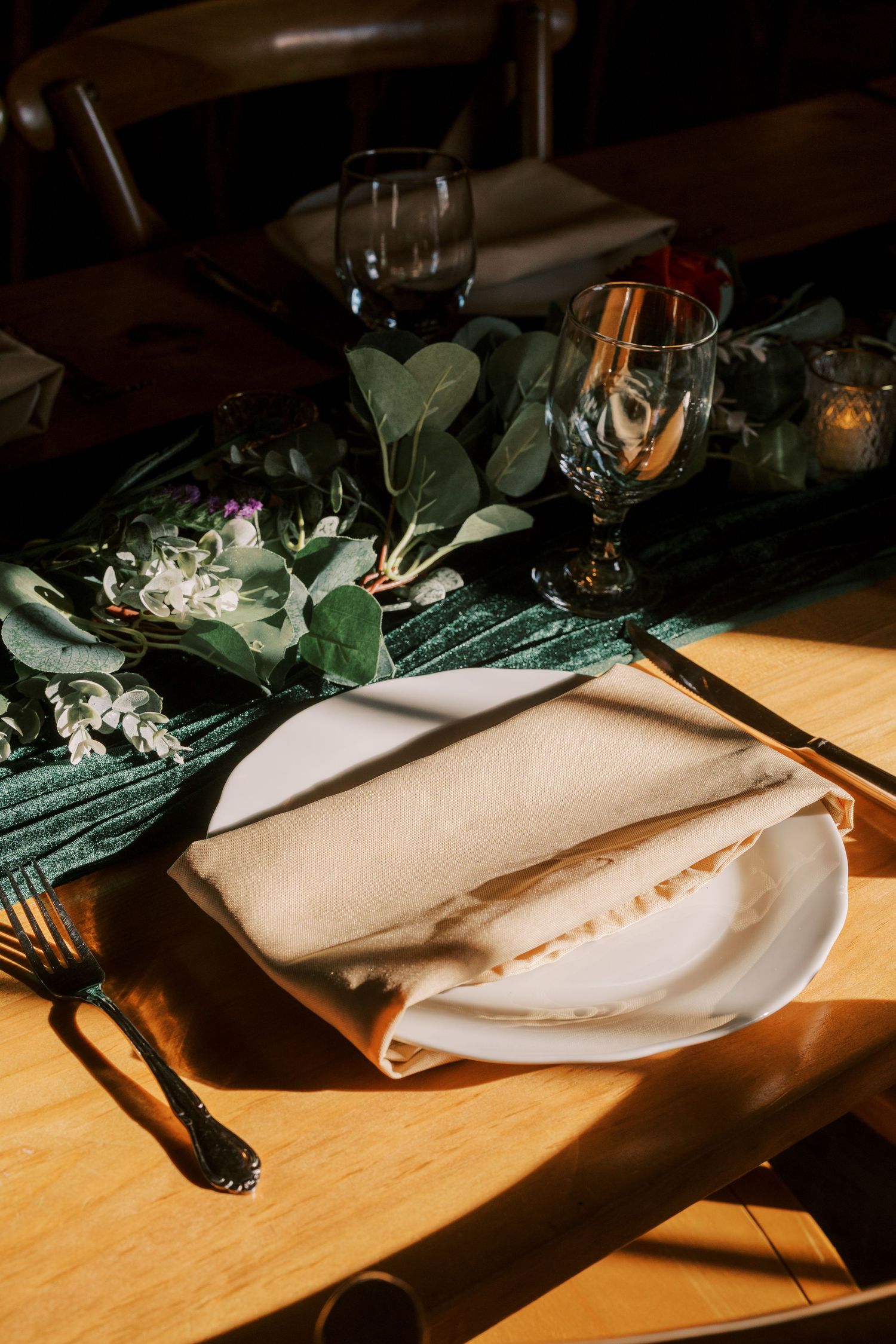 Place settings at a restaurant table feature white napkins, green foliage, and dramatic lighting on a wooden surface.