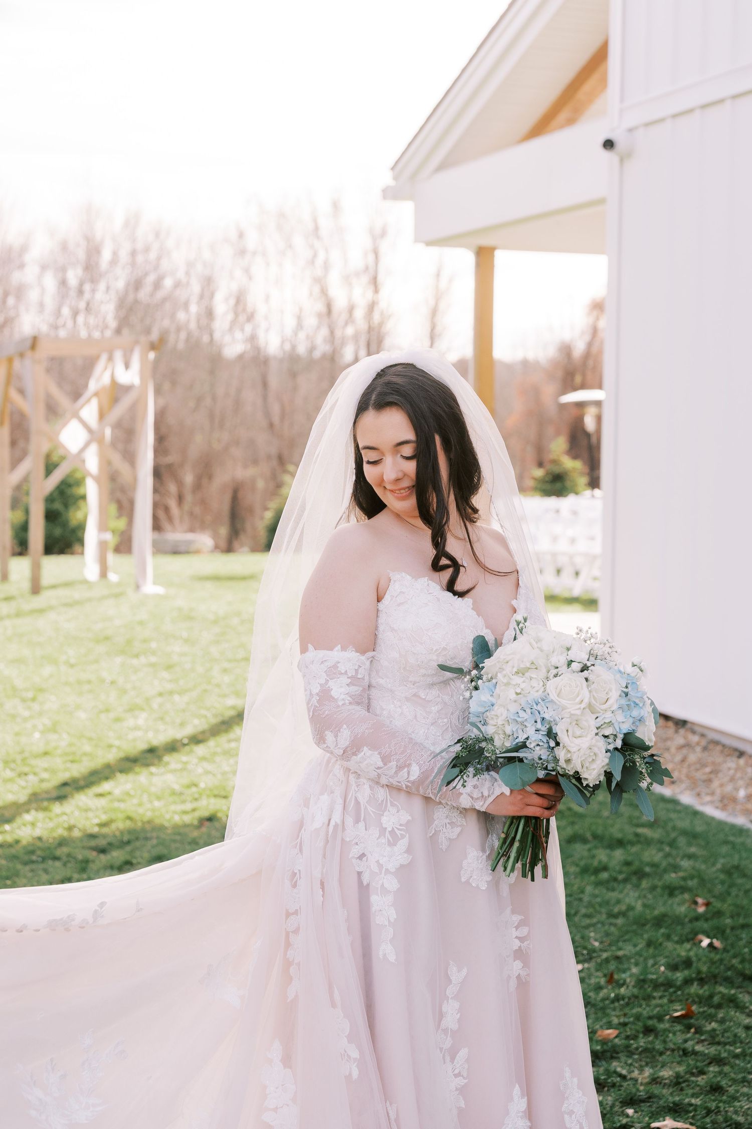 A bride in a flowing white lace gown poses outdoors at a white gazebo during a romantic garden wedding photoshoot.