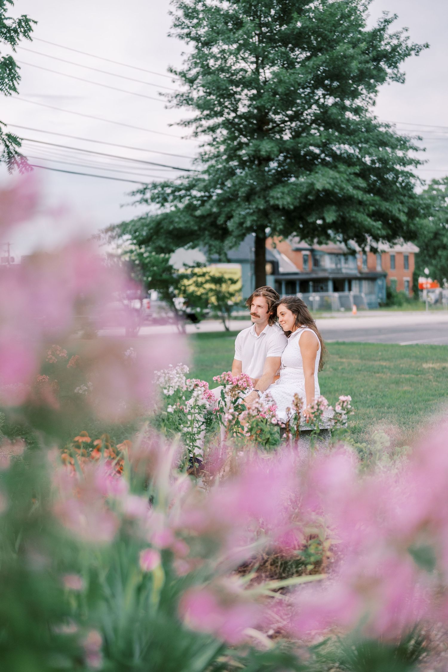 Couple embraces in a garden with pink flowers blooming in the foreground against green foliage.