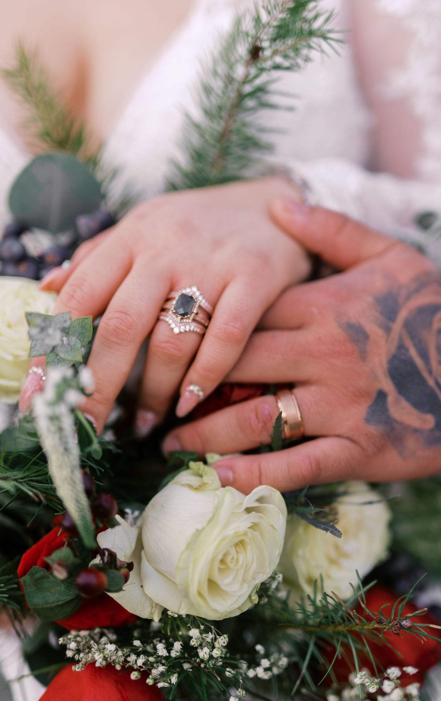 Wedding hands with rings and tattoos rest on a bouquet of white roses and evergreen sprigs.