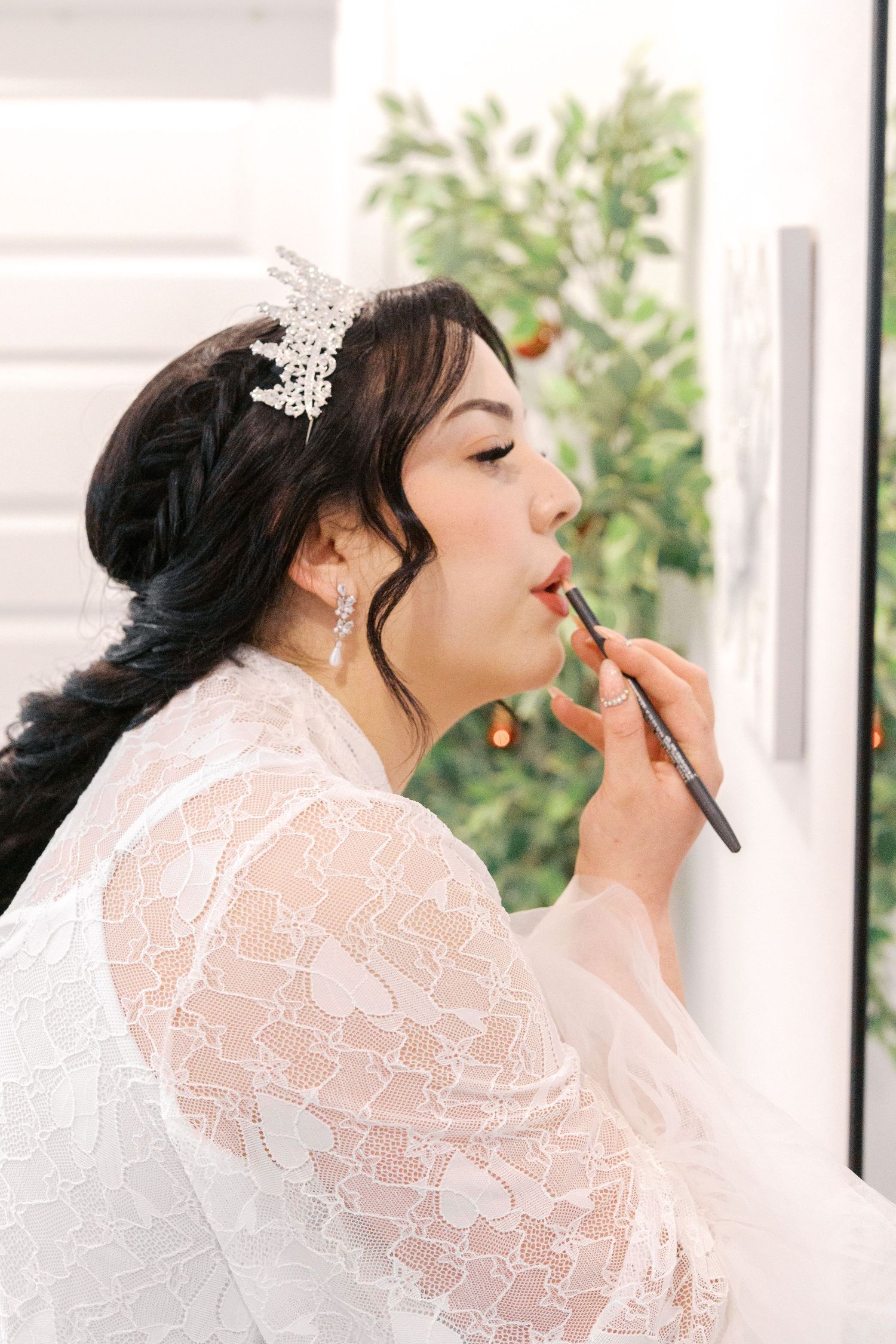 A bride in a lace robe getting ready for her wedding day, applying lipstick while wearing a crystal hair accessory.