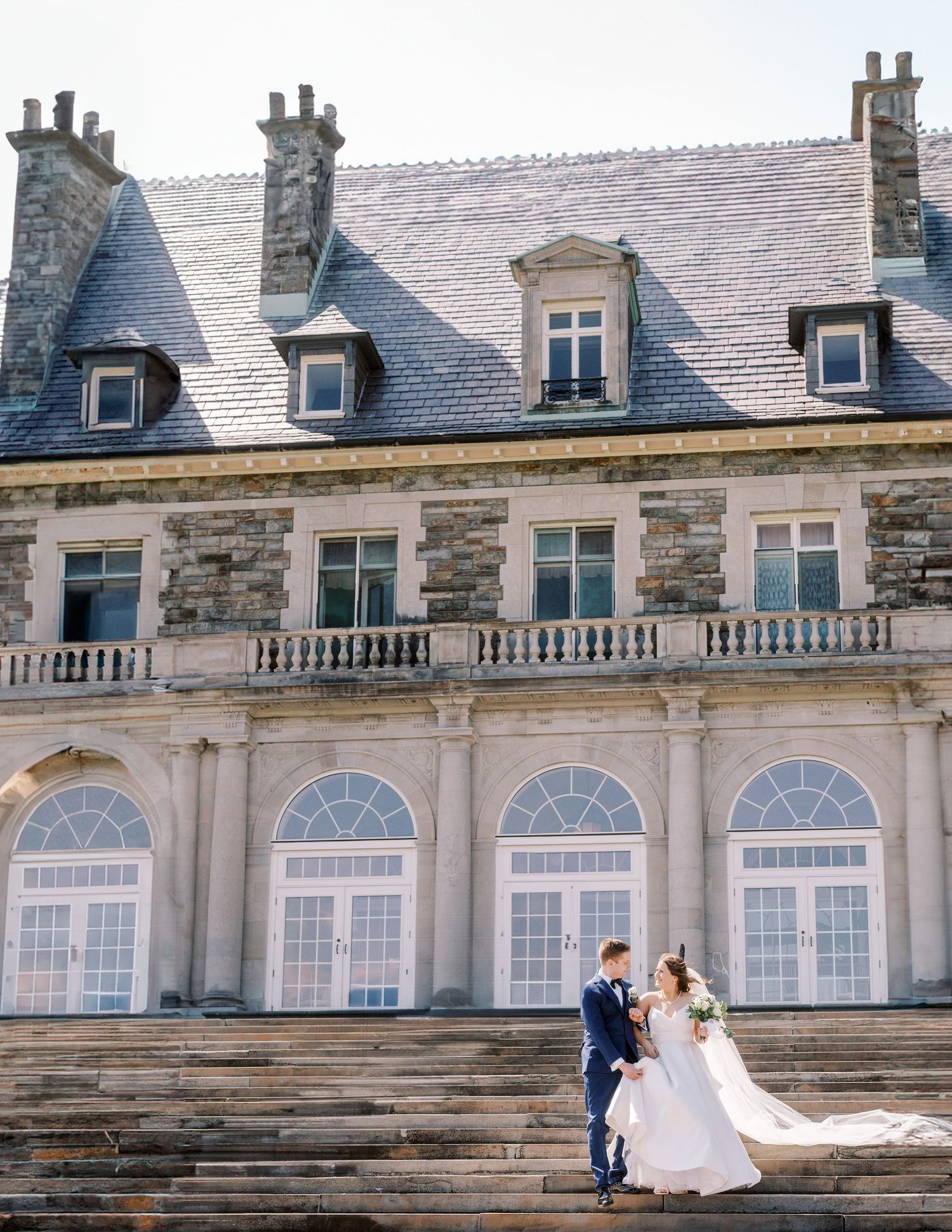 A couple poses on stone steps in front of a grand historic mansion with arched windows and balustrades.