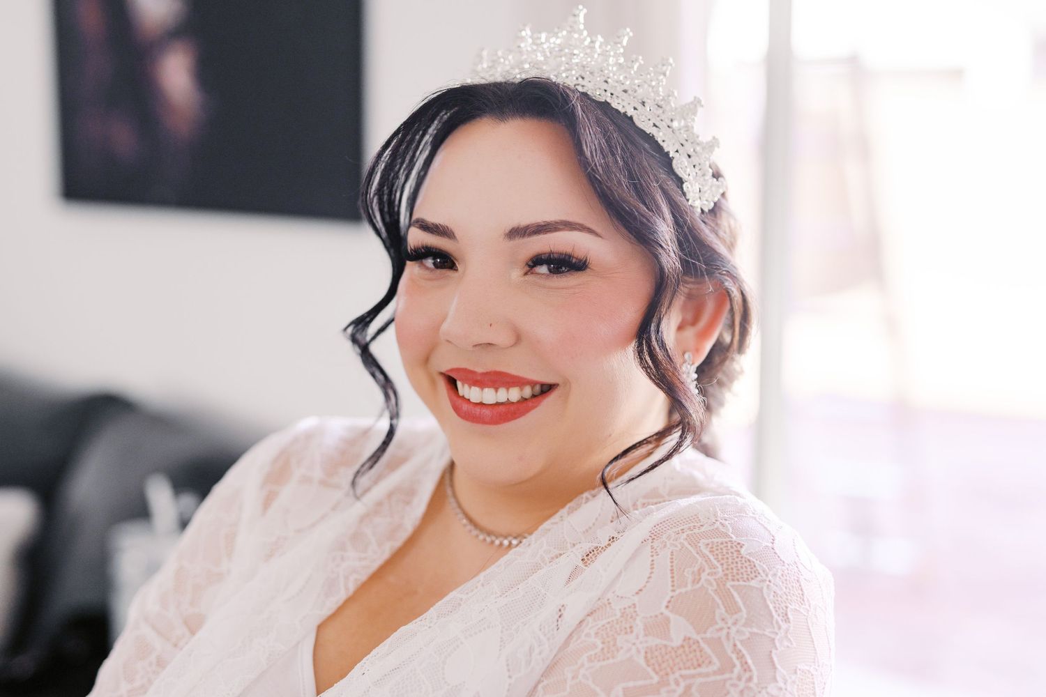 A bride in white lace smiles warmly while wearing a delicate crystal tiara and red lipstick.