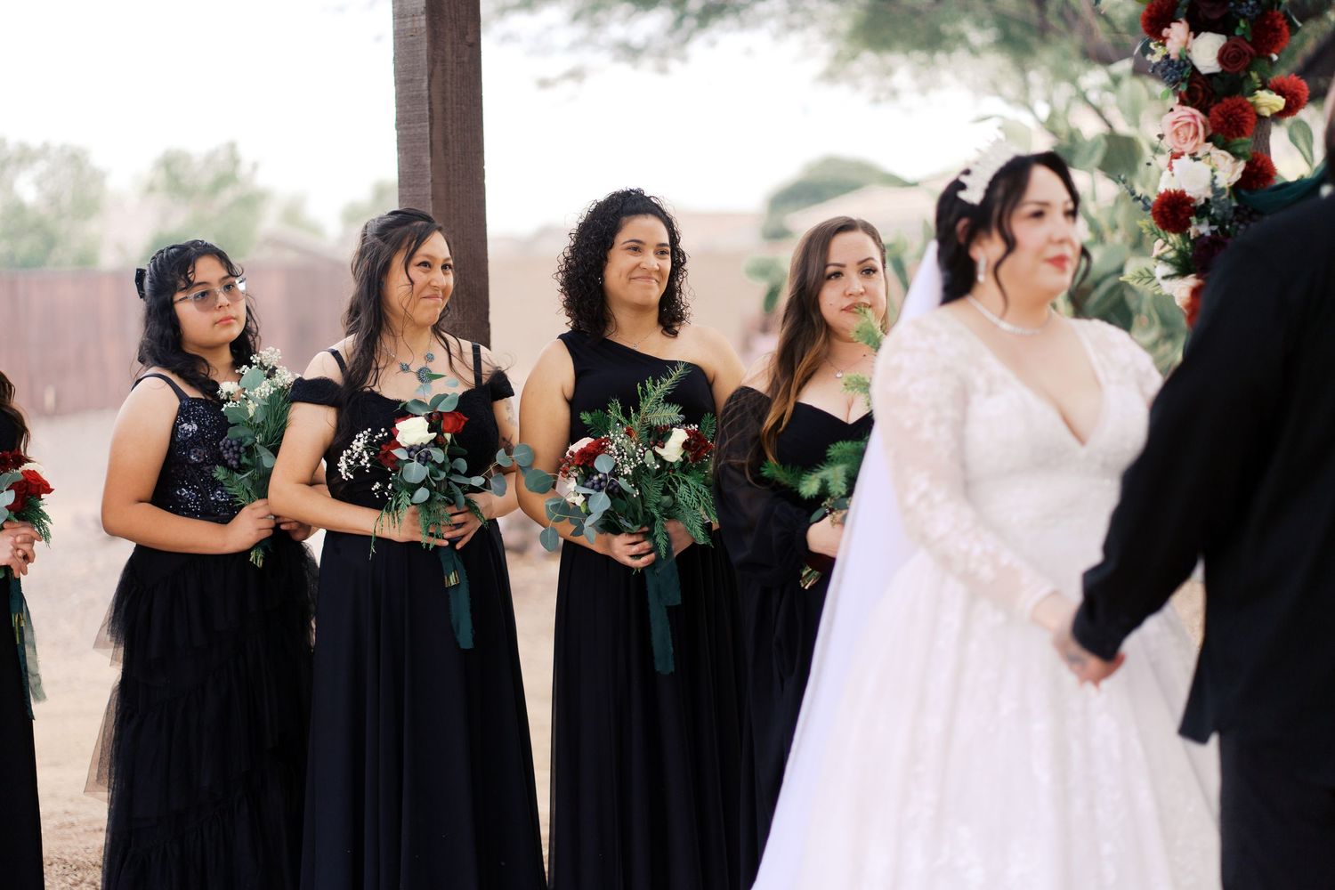 Bridesmaids in elegant black dresses holding bouquets stand beside bride in white gown during outdoor wedding ceremony.