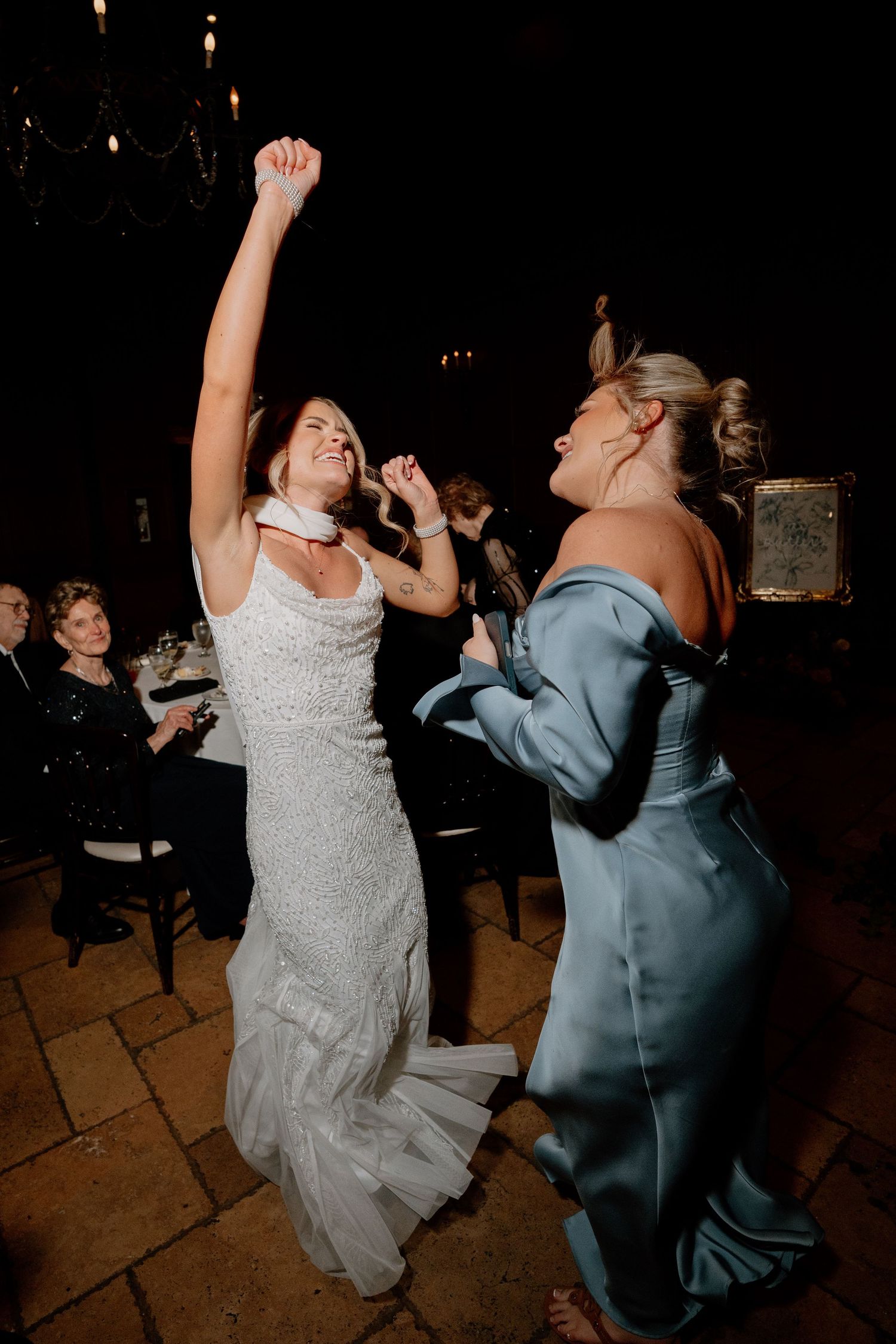 Two wedding guests dance energetically on a dark dance floor with one wearing a white dress and another in blue attire.