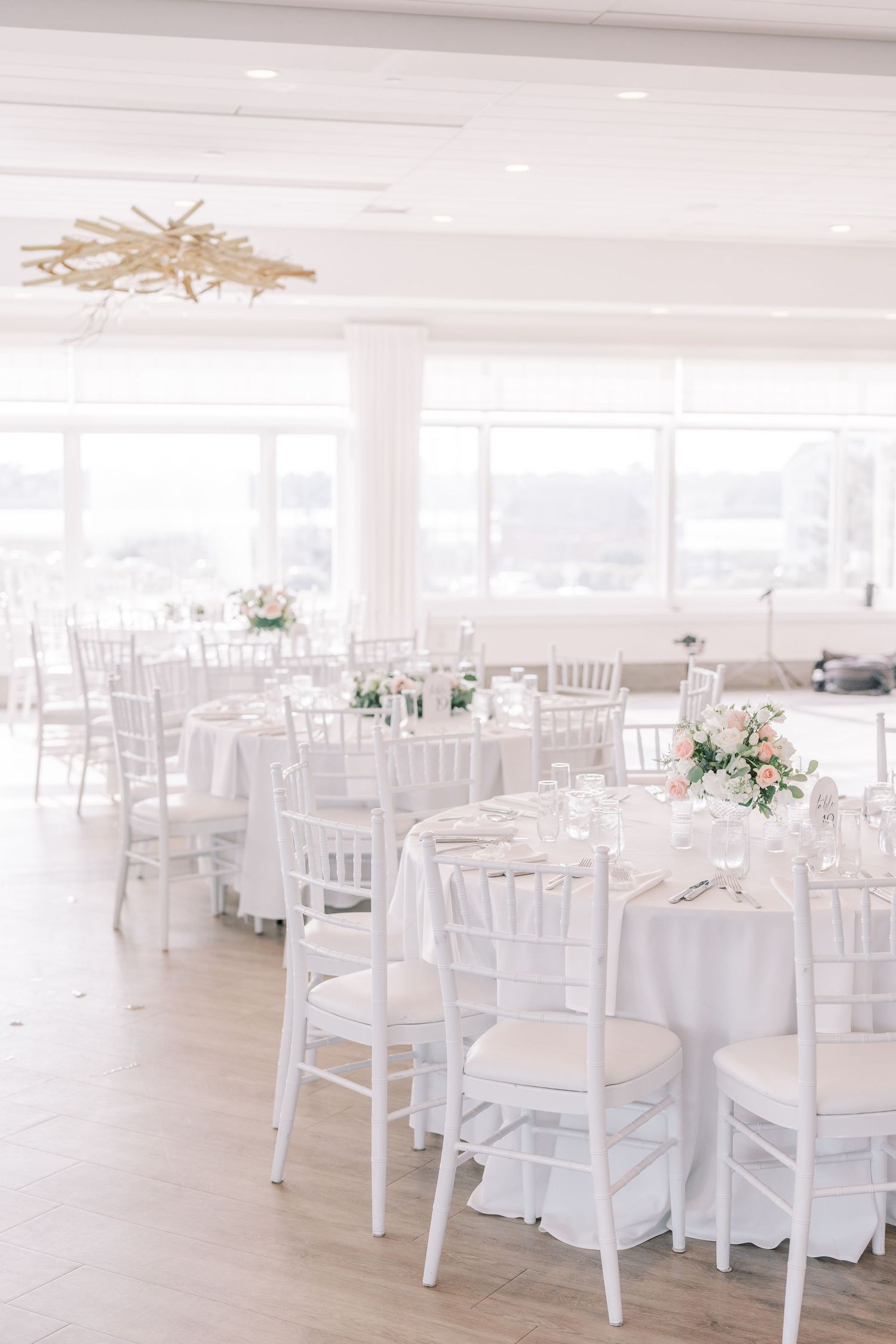 White wedding reception tables and chairs arranged in an elegant ballroom with soft natural lighting and floral centerpieces.