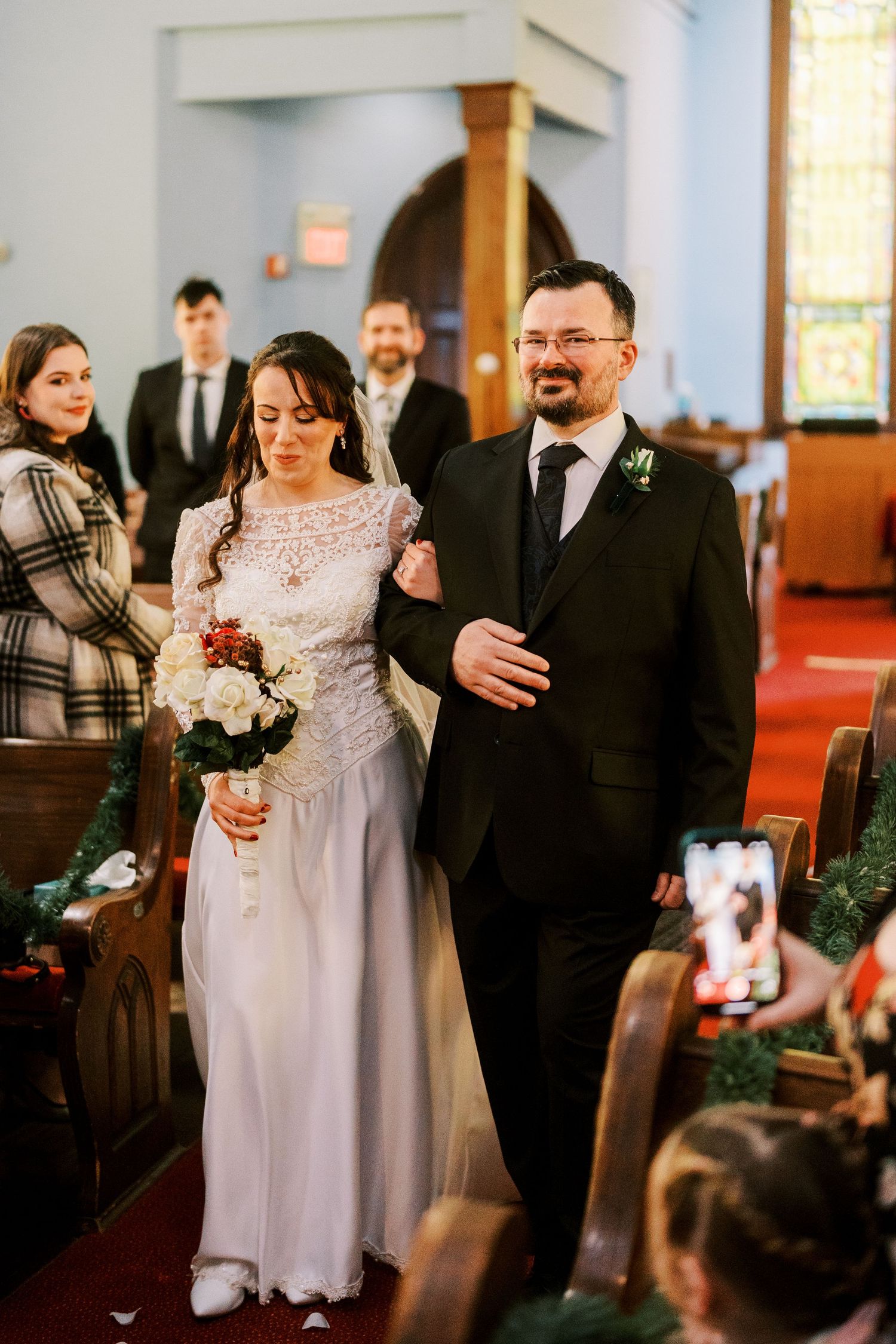 Wedding guests gather in a church with red carpeting as they capture photos on their phones.