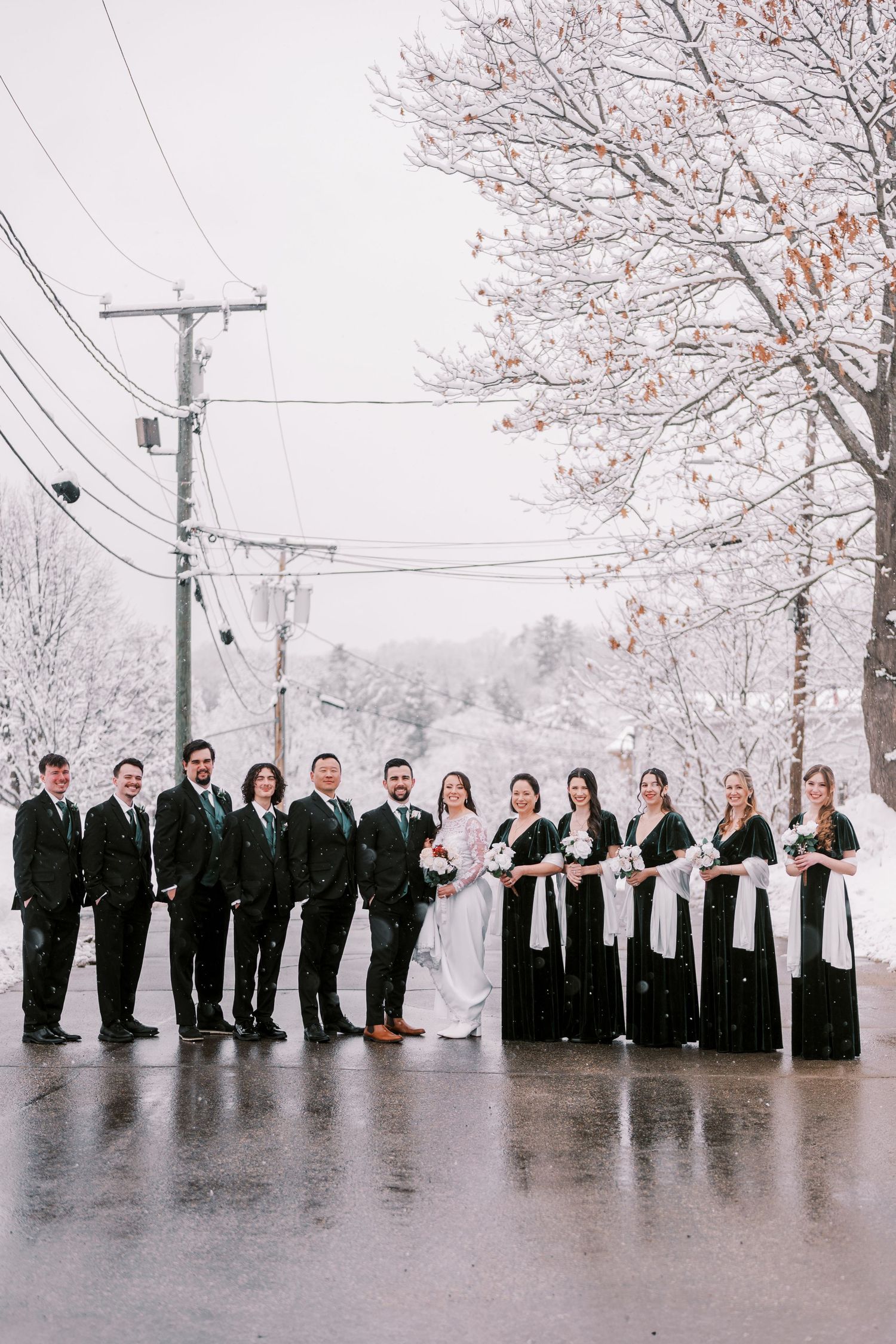 Wedding party lined up outdoors in winter with snow covered trees and utility poles in background.