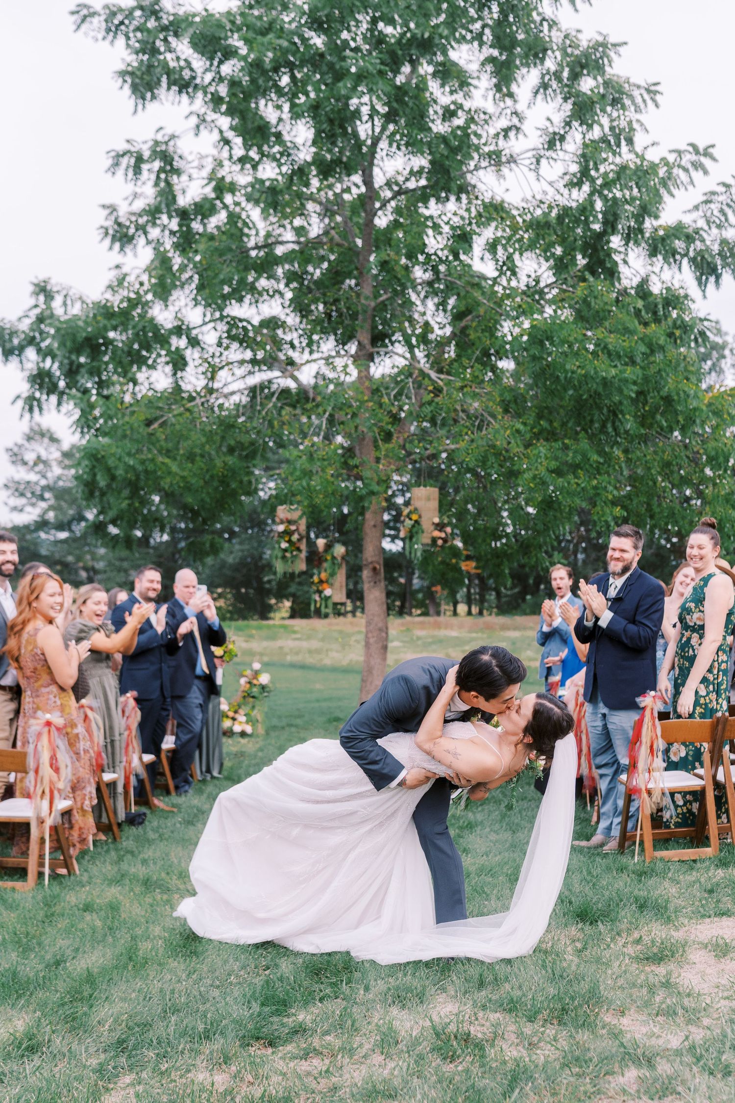Wedding ceremony setup with wooden chairs along tree-lined aisle in rustic outdoor setting.