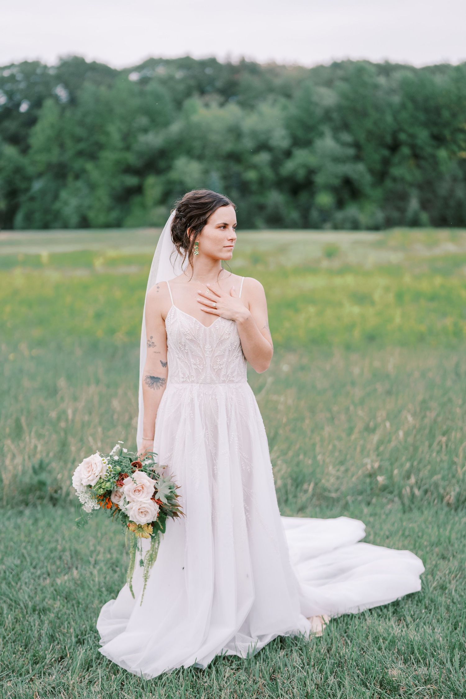 Bride holds bouquet while posing in delicate white gown with long train in summer field.