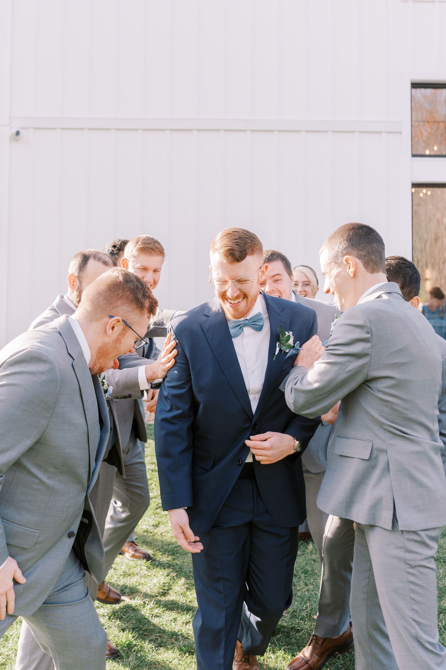 Wedding party members in navy blue and gray suits share a candid laughing moment together outdoors.