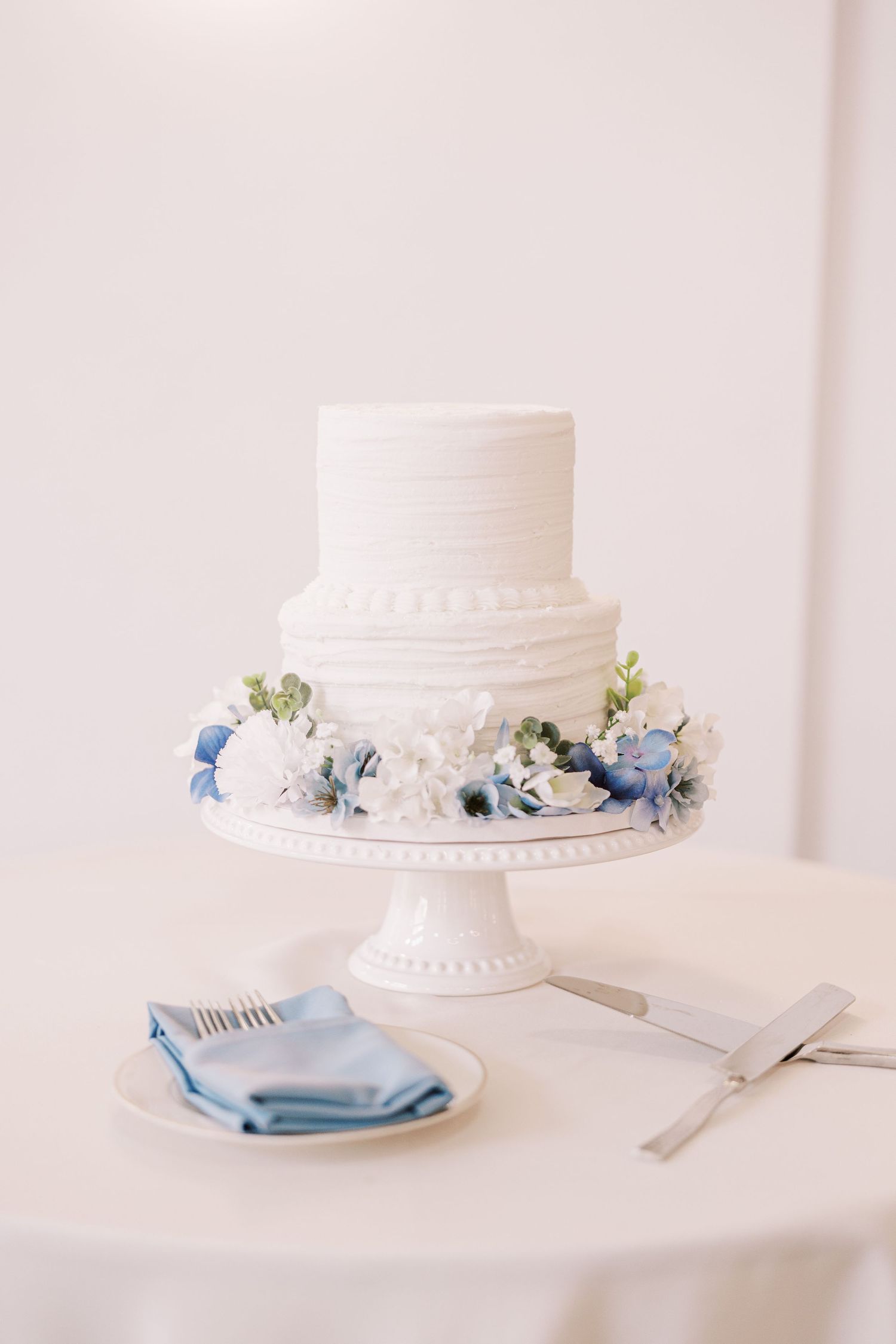 Elegant white two-tier wedding cake decorated with blue and white flowers on a classic pedestal stand with table settings.