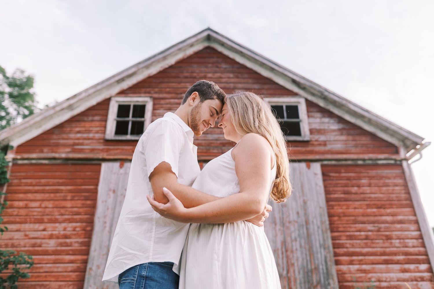 A romantic couple embraces in front of a rustic red barn during a summer wedding photoshoot.