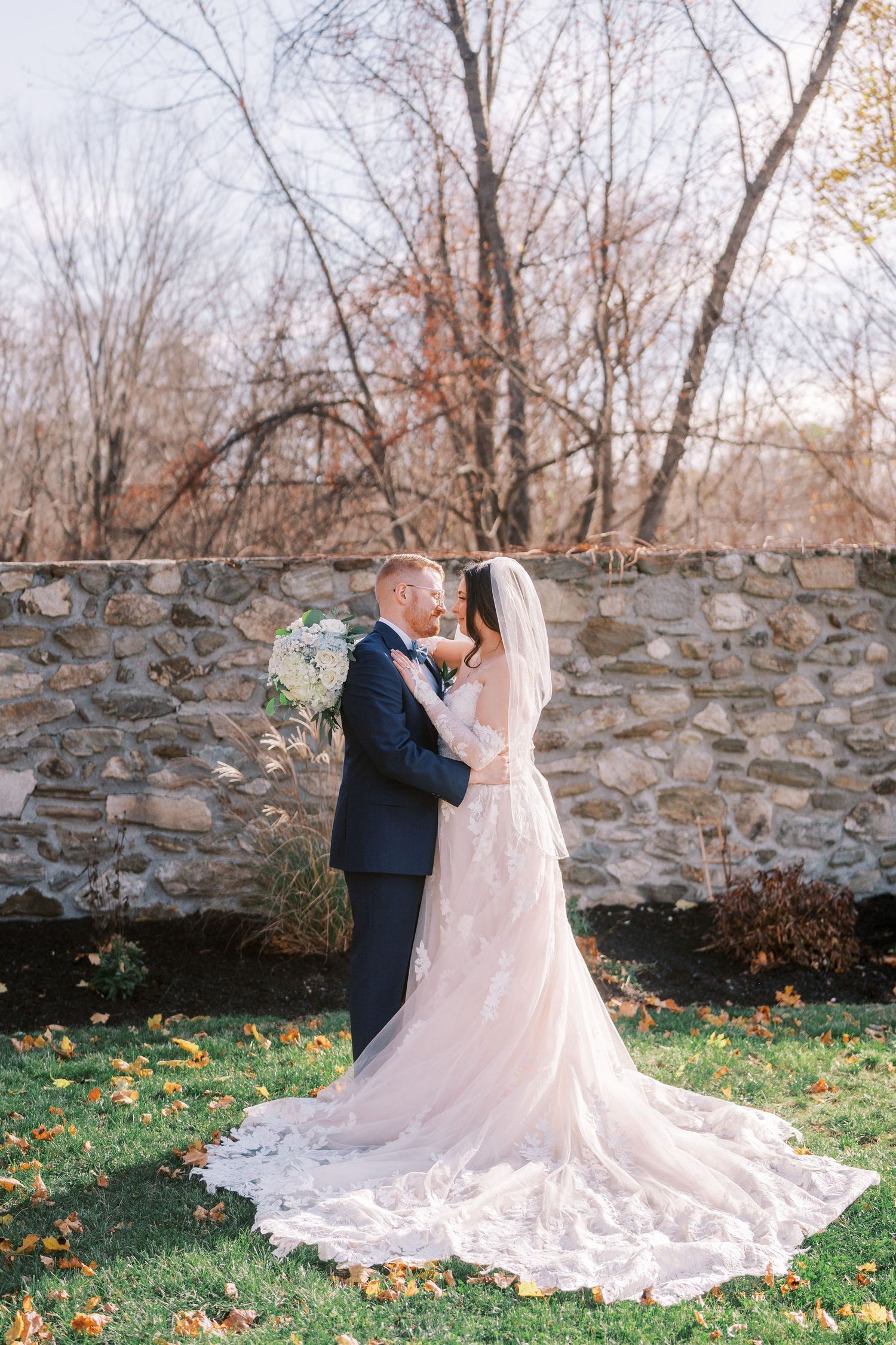 A couple embraces in front of a rustic stone wall during an autumn wedding, with a flowing lace train on display.