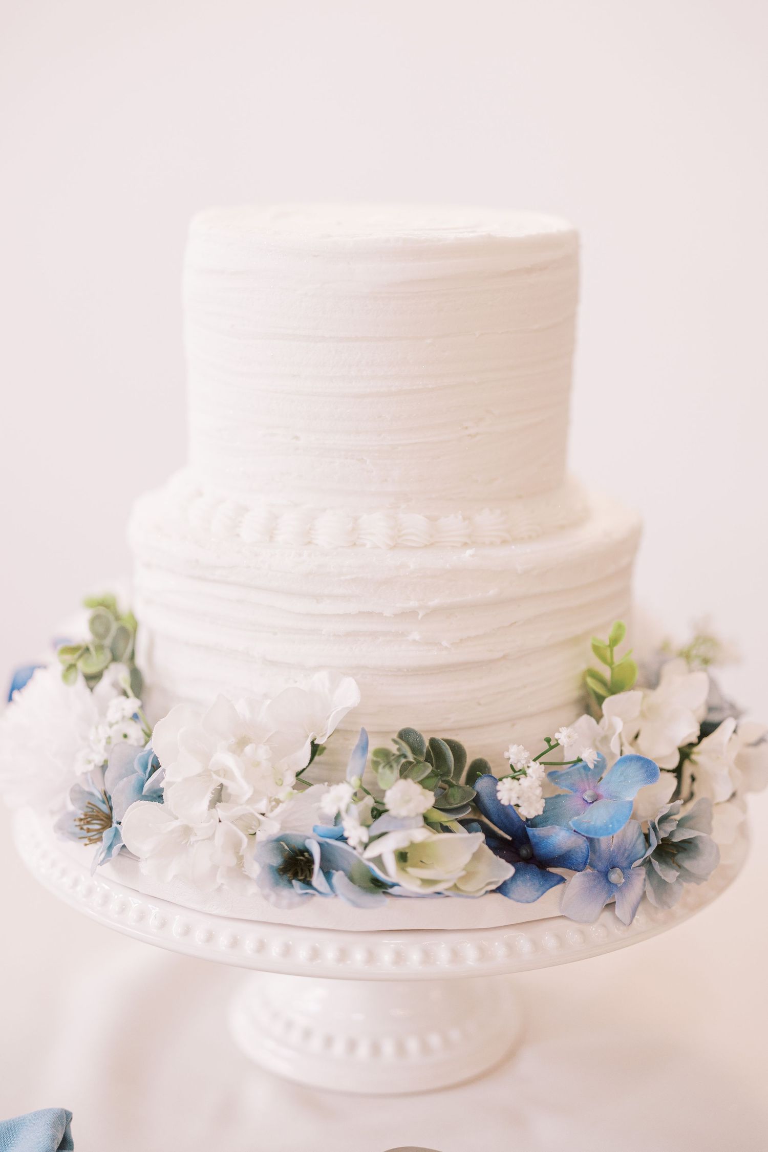 Two-tiered white wedding cake with textured frosting and delicate blue and white floral decorations on a white pedestal stand.