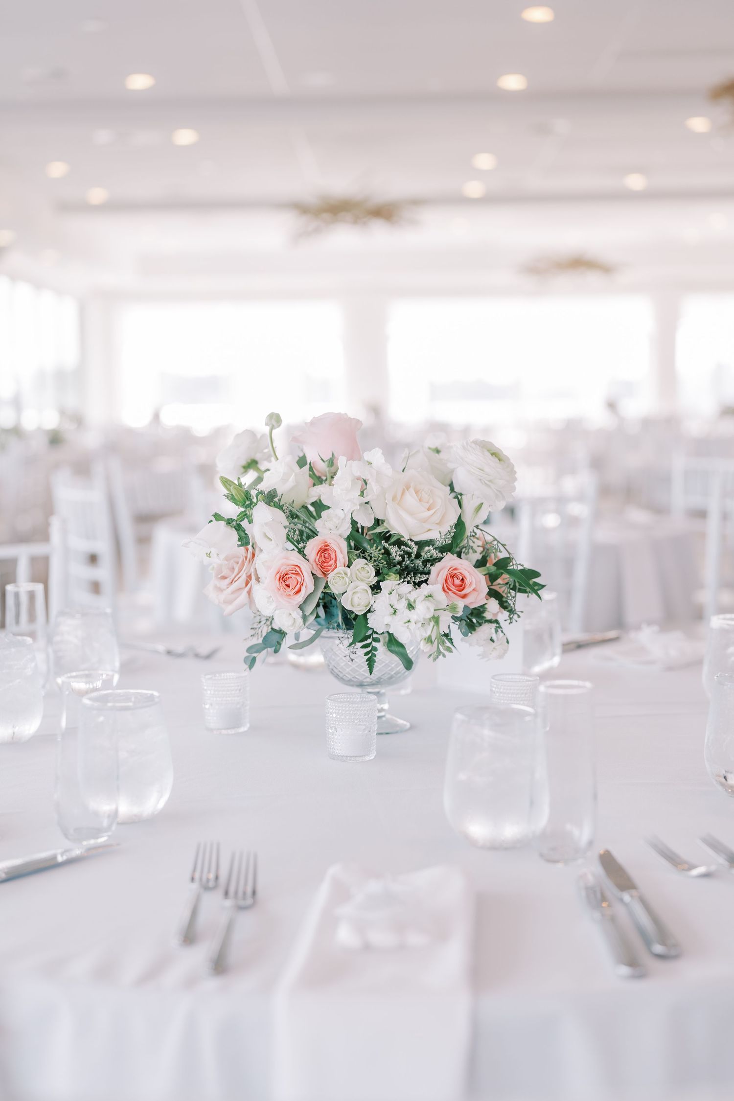 Elegant white wedding reception tables with soft pink and white floral centerpieces and crystal glassware in bright venue.