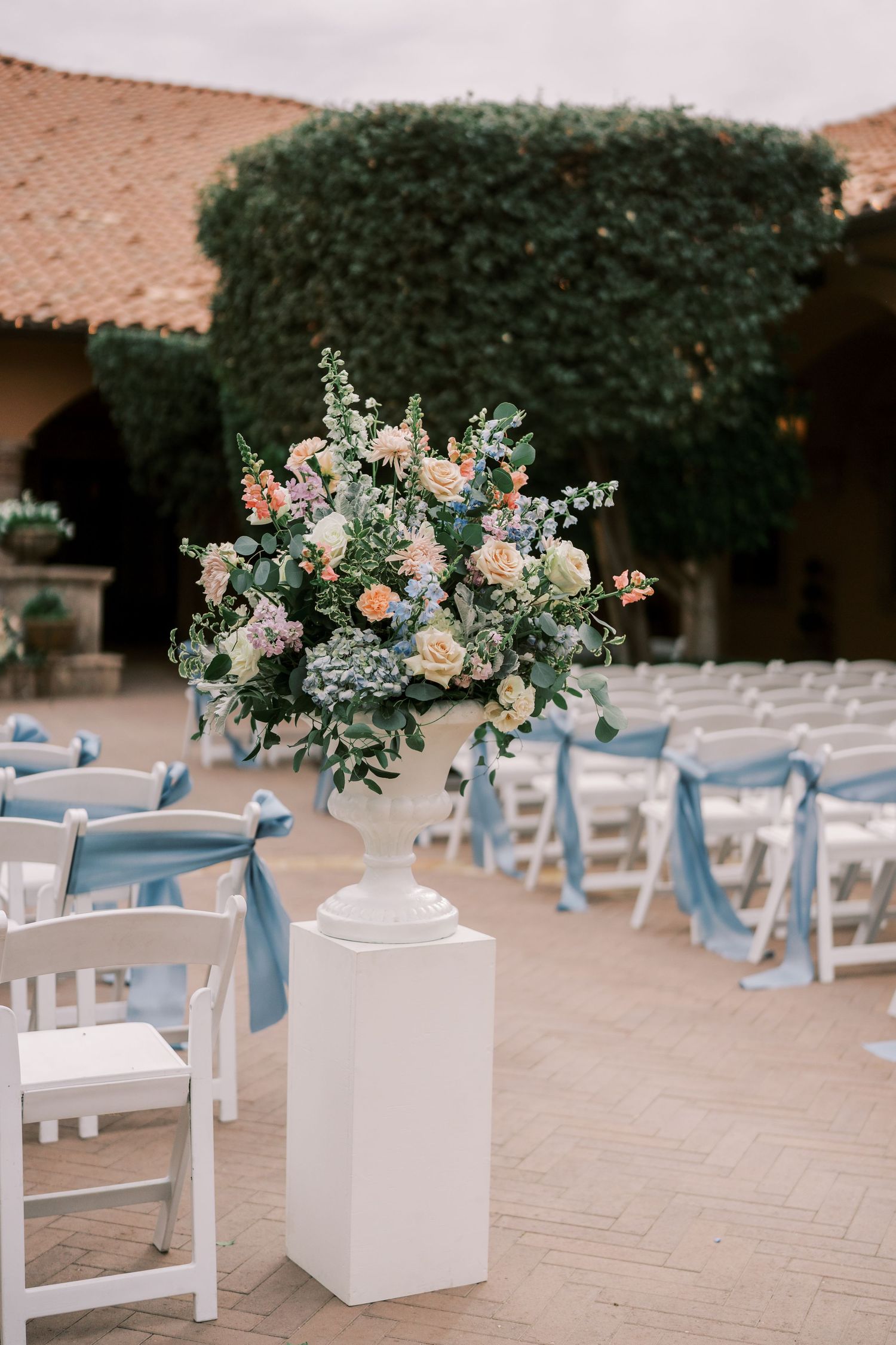 Outdoor wedding ceremony setup with white chairs adorned with light blue ribbons and floral arrangements on white pedestals.