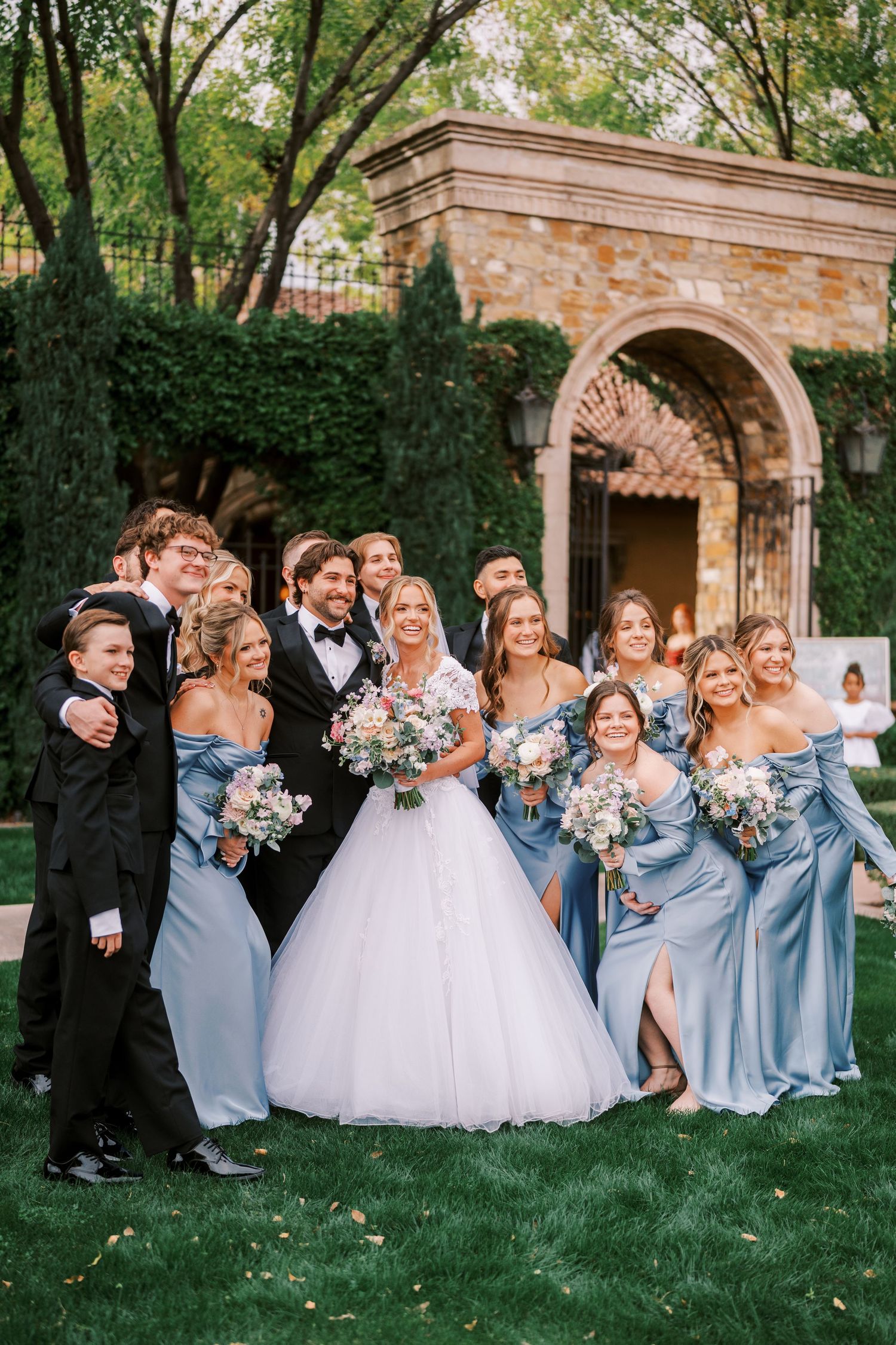 Wedding party in light blue bridesmaid dresses and black tuxedos pose together on grass near stone archway.
