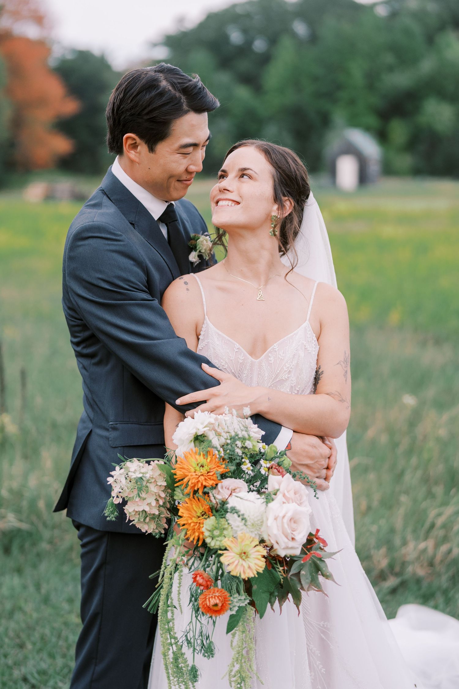 A newlywed couple embraces in a meadow with the bride holding a colorful summer bouquet.