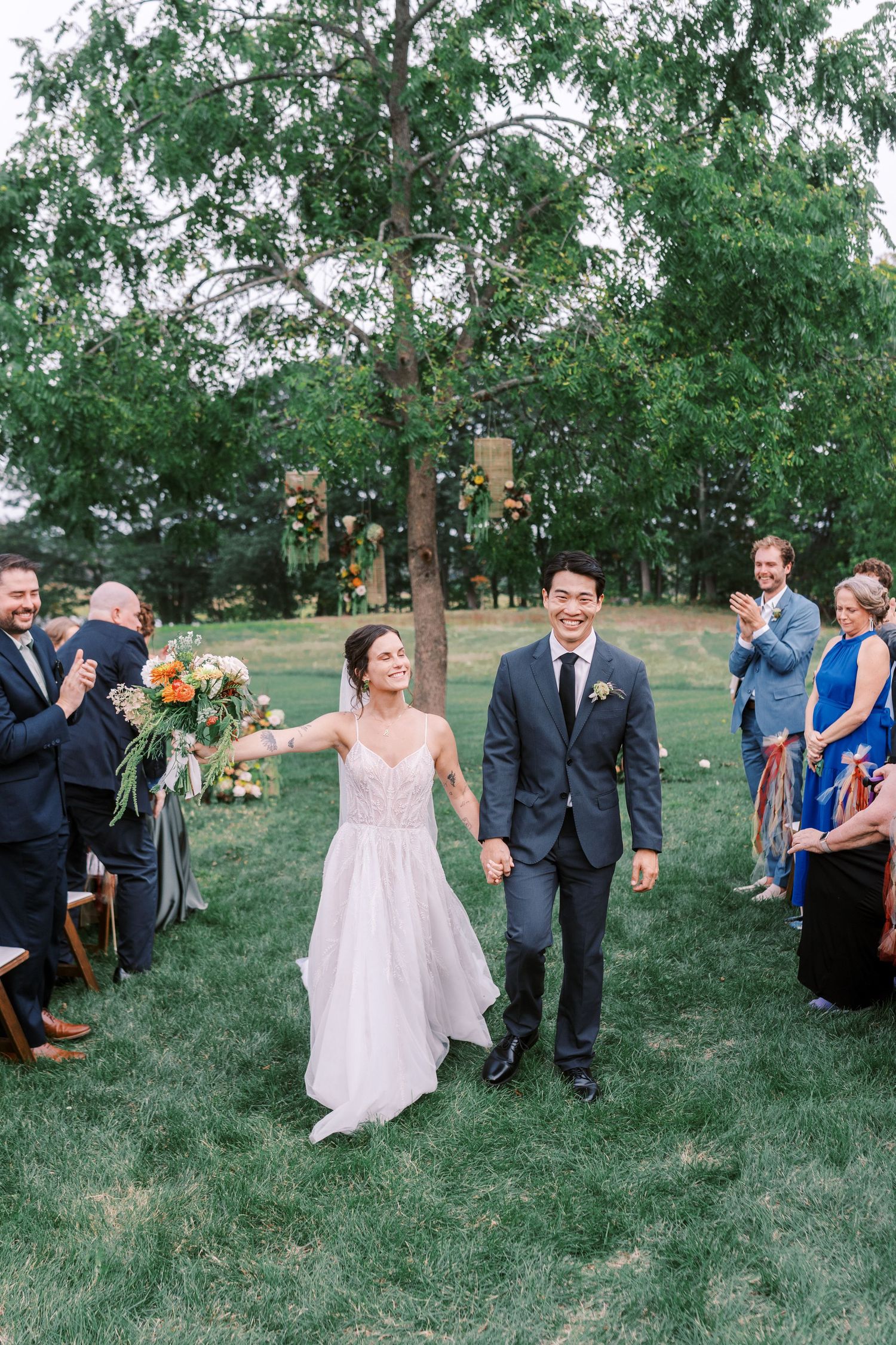 Wedding party celebrates outdoors under trees with decorative lanterns hanging from branches.