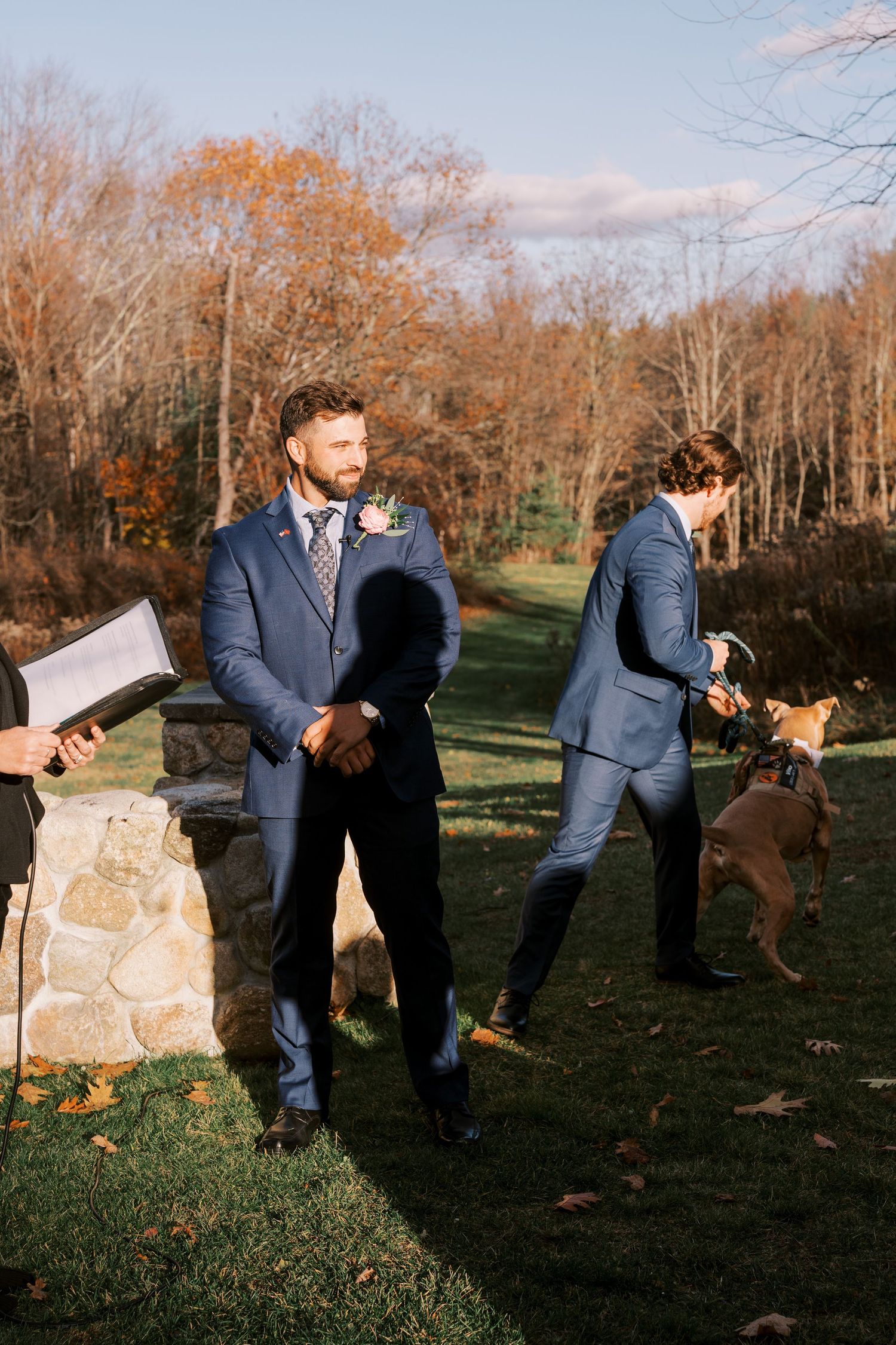 A wedding party in blue suits enjoys a sunny autumn day outdoors with their dogs during a celebration.