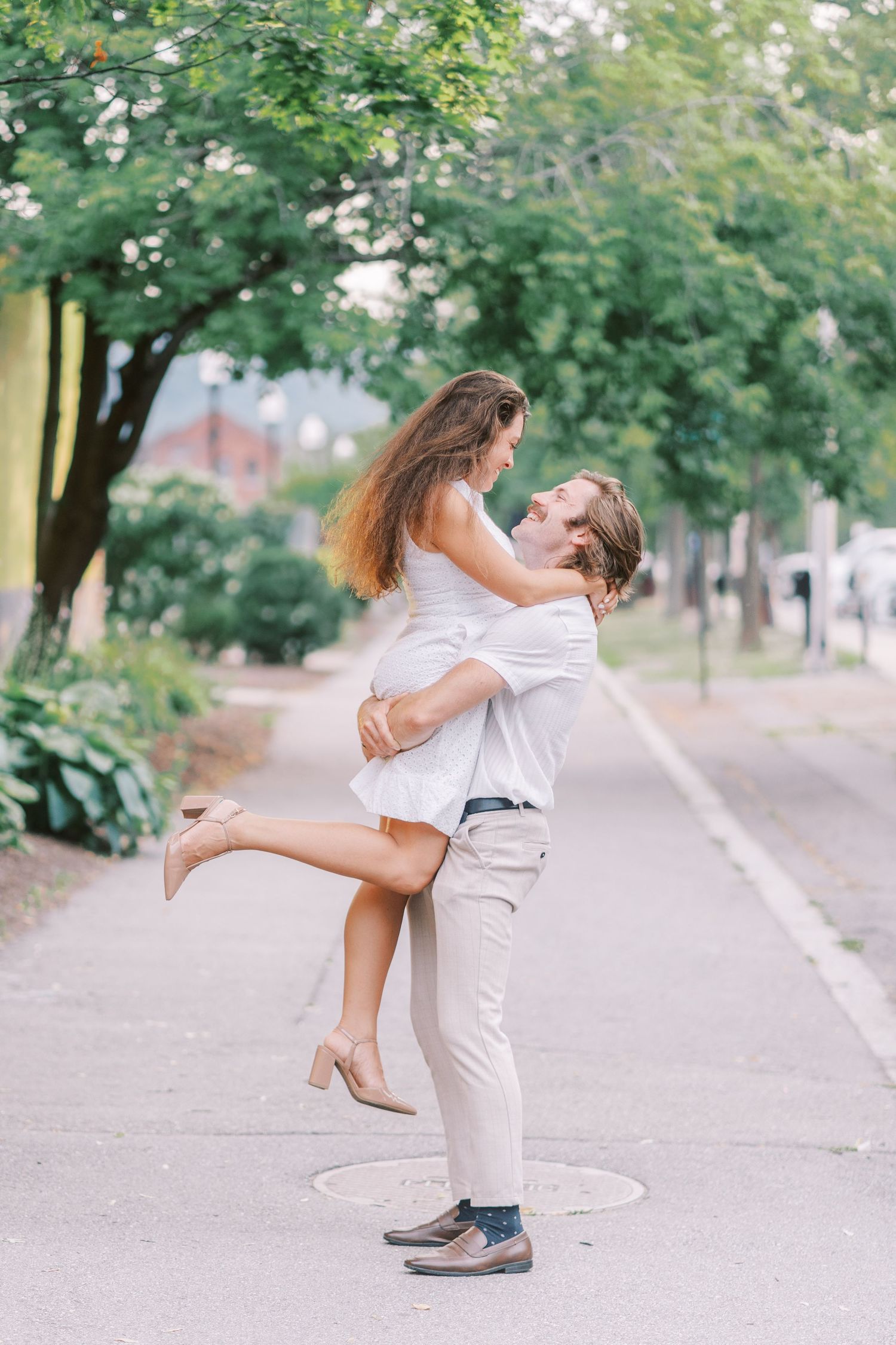 A couple in white clothing spins and dances together on a tree-lined path.