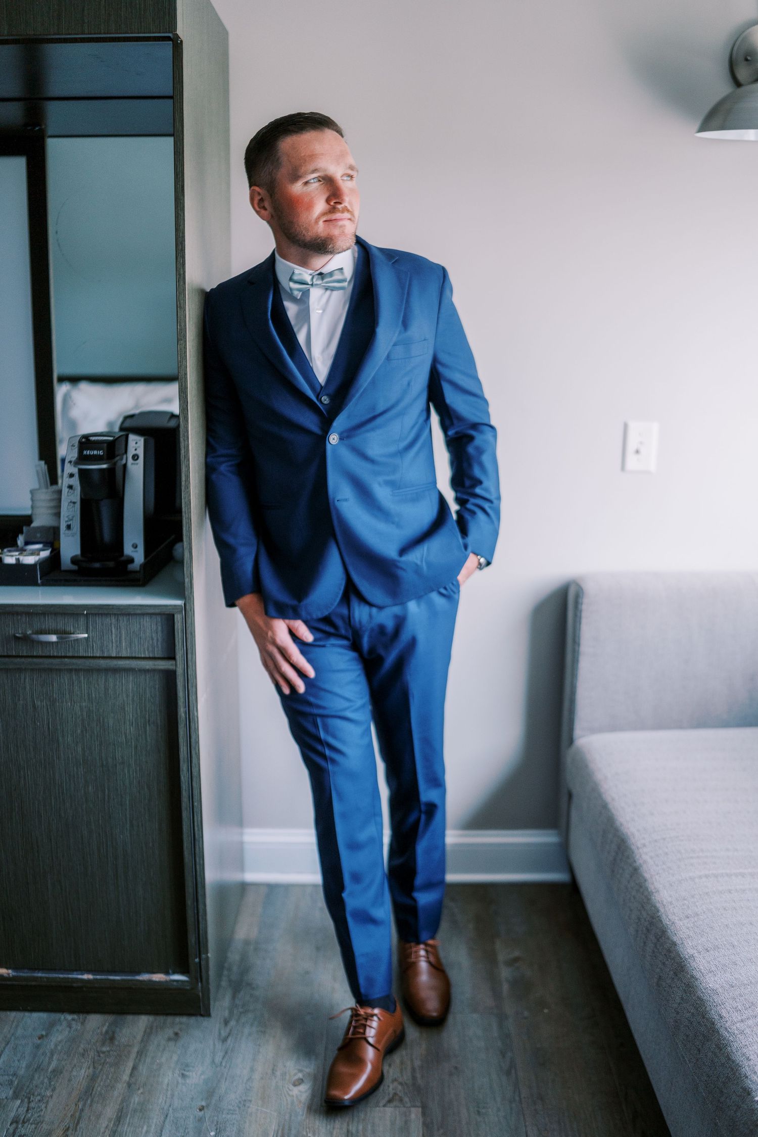 A person in a tailored blue suit and brown dress shoes poses by a modern gray sofa in an interior setting.