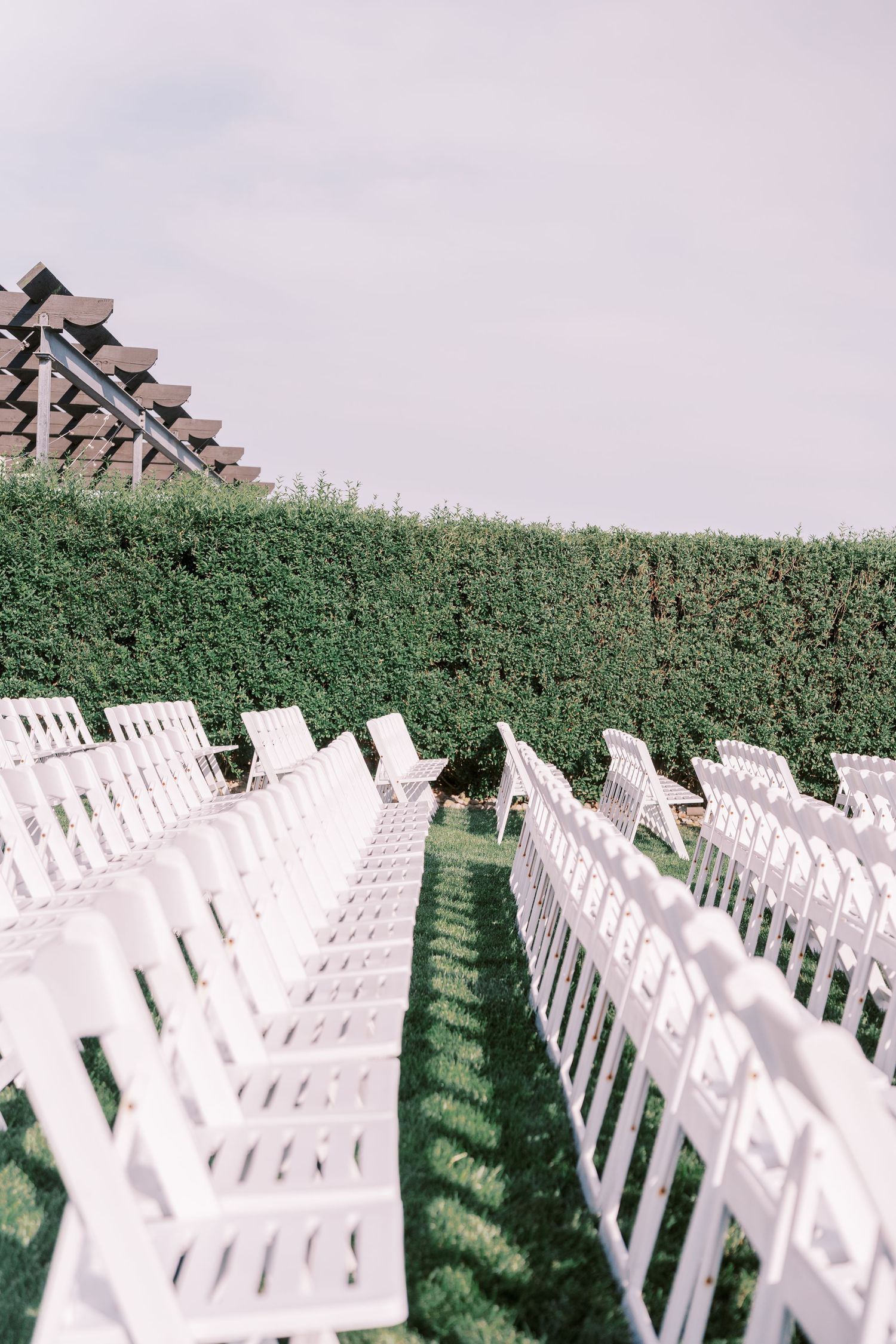 Rows of white folding chairs arranged on a lawn with green hedges and rustic buildings in the background for an outdoor event.