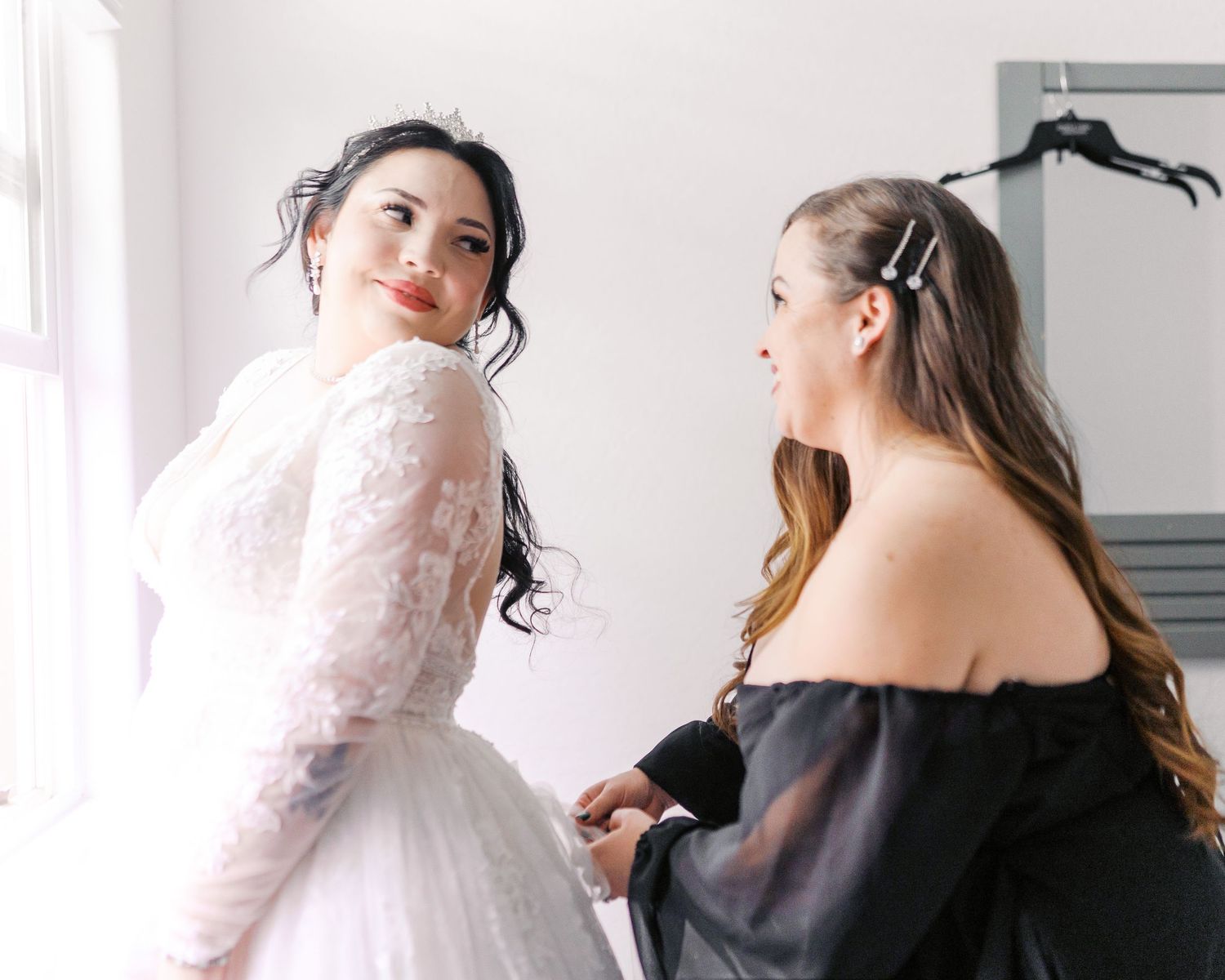 Two women in wedding attire share a tender moment while getting ready in a bright room.