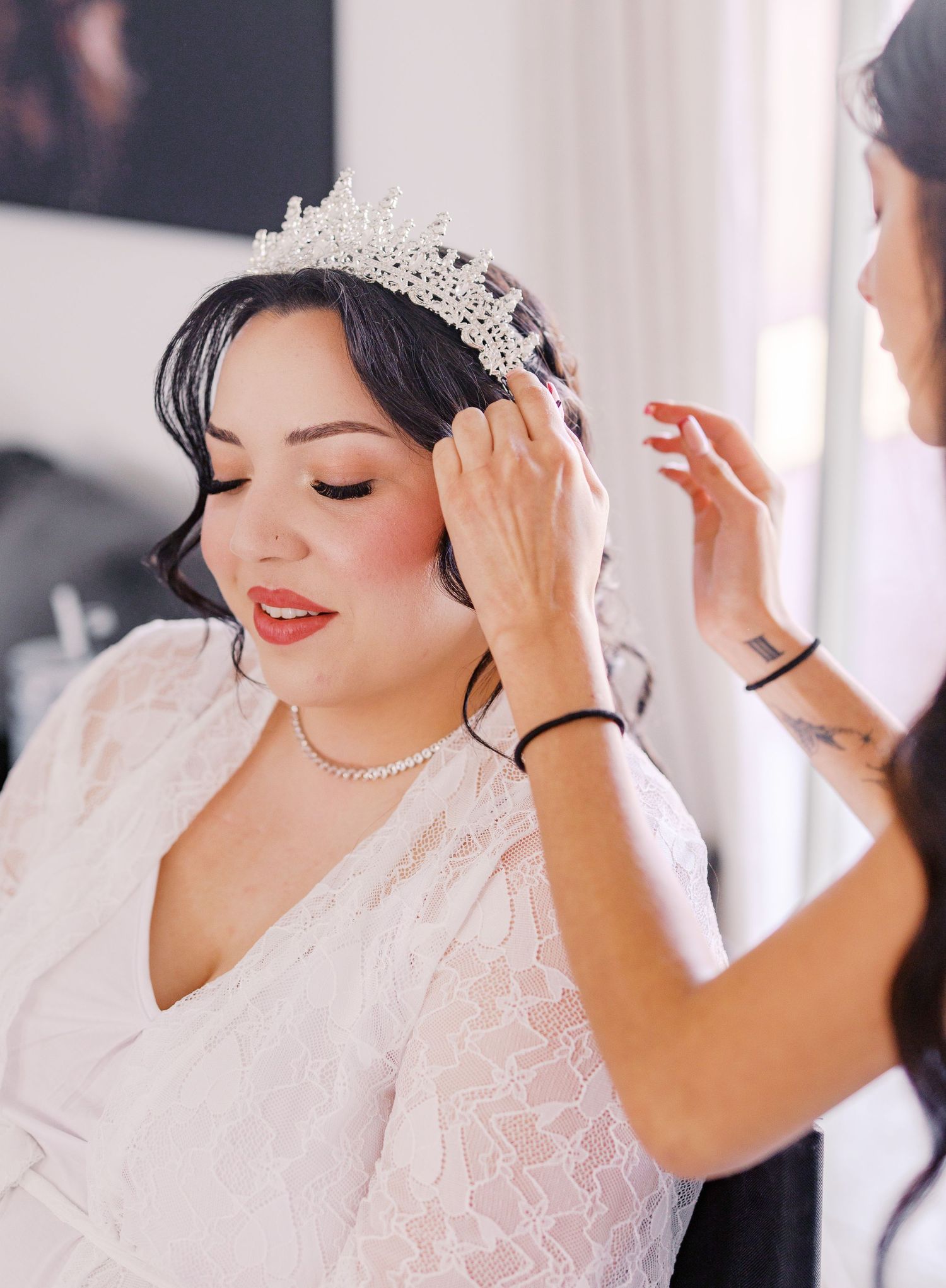 A bride in a white lace dress and sparkly tiara gets her final styling touches done before her wedding ceremony.