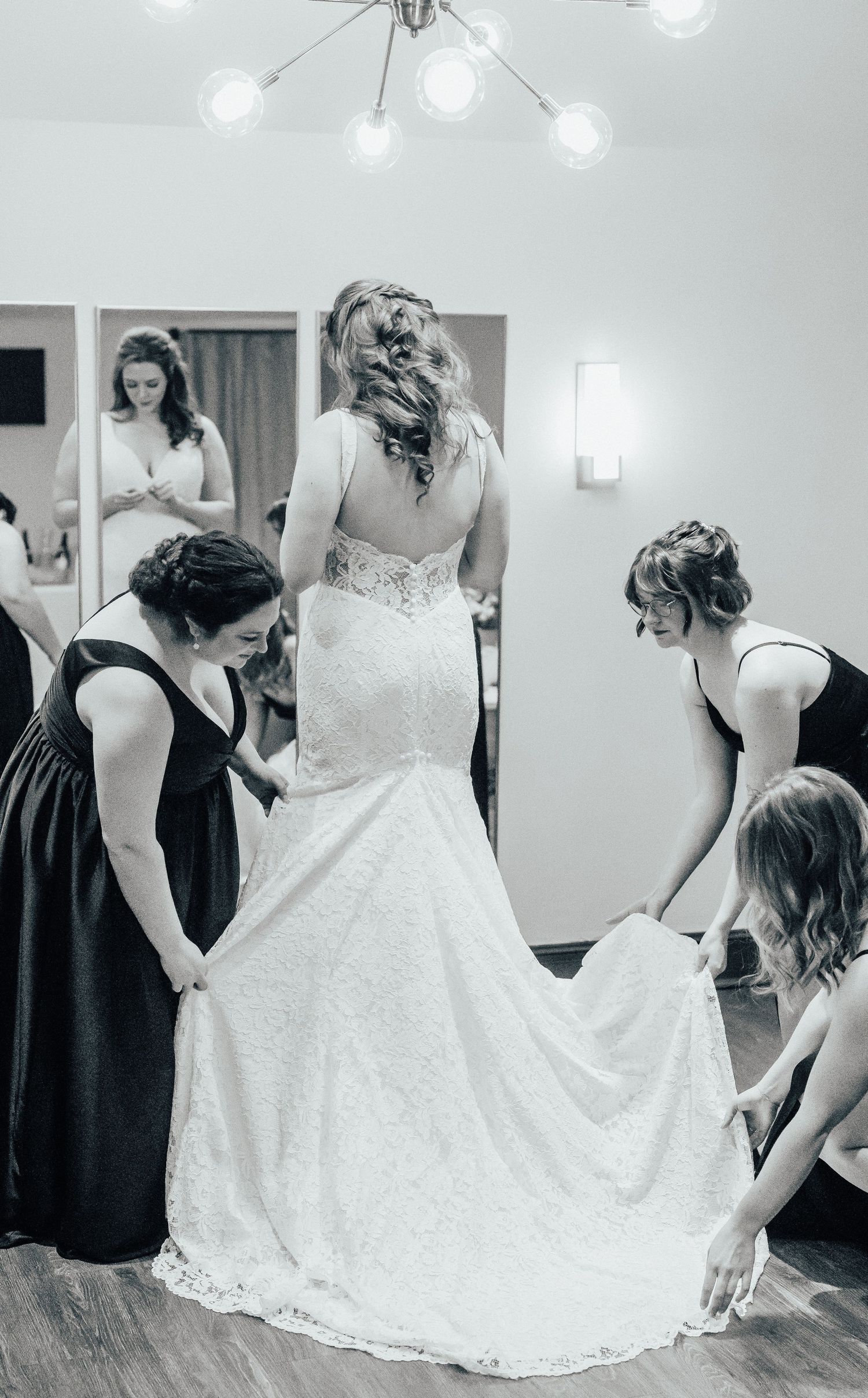 Bridesmaids helping adjust a beautiful lace wedding gown in a dressing room.