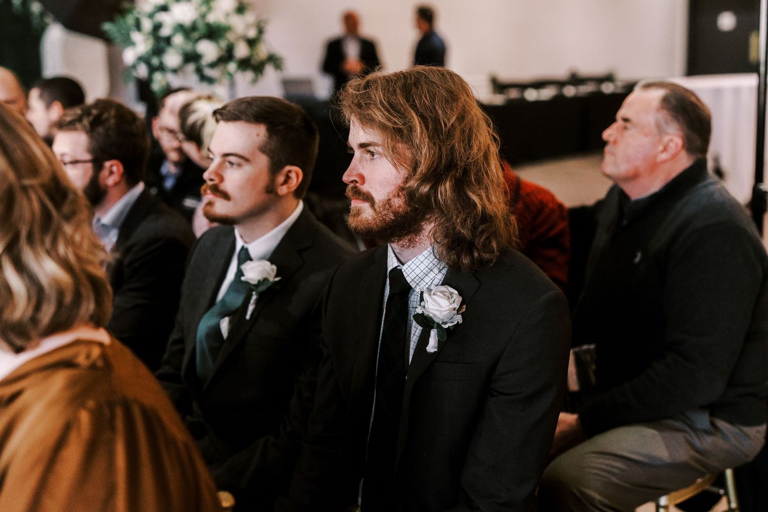 Wedding guests in formal attire seated during an indoor ceremony with floral decor visible in background.