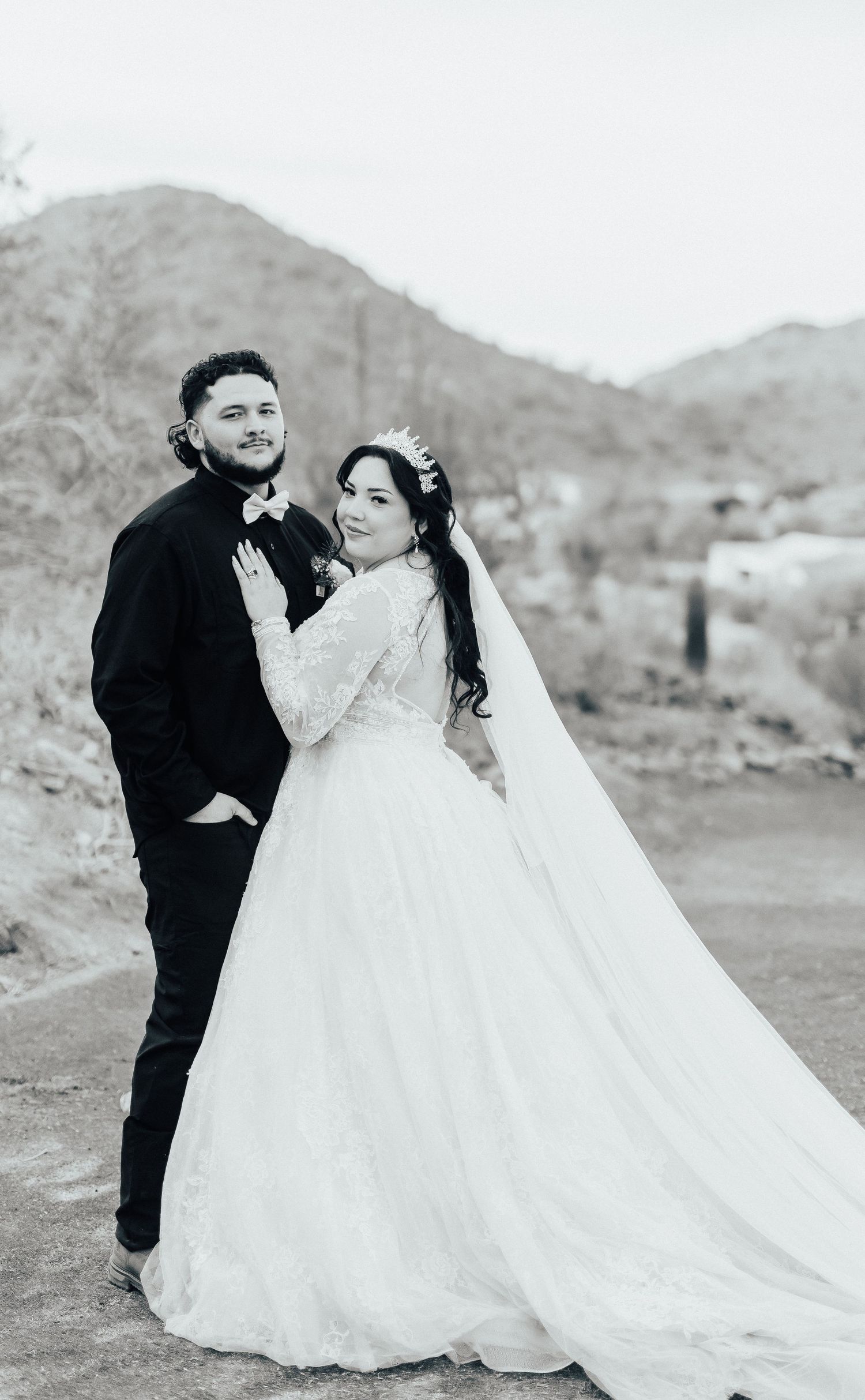A couple in wedding attire poses together against a desert mountain landscape in black and white photography.