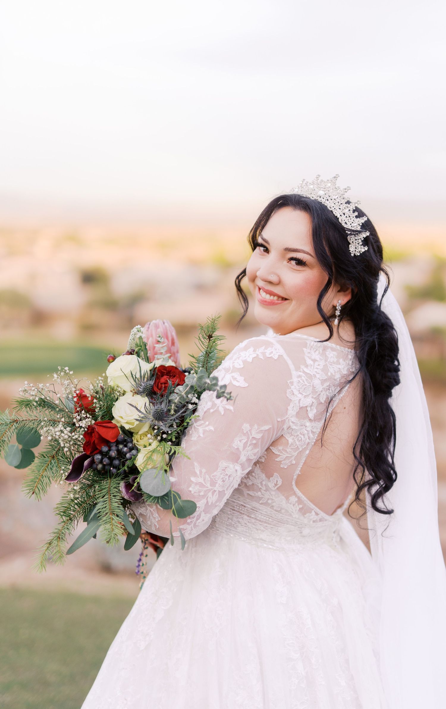 A stunning bride in a white lace gown with long sleeves holds a colorful bouquet while posing outdoors at sunset.