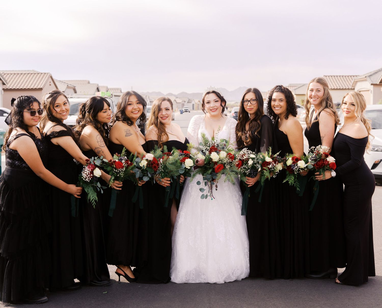 Bridal party in black dresses holding burgundy and white floral bouquets pose together outdoors on rooftop.