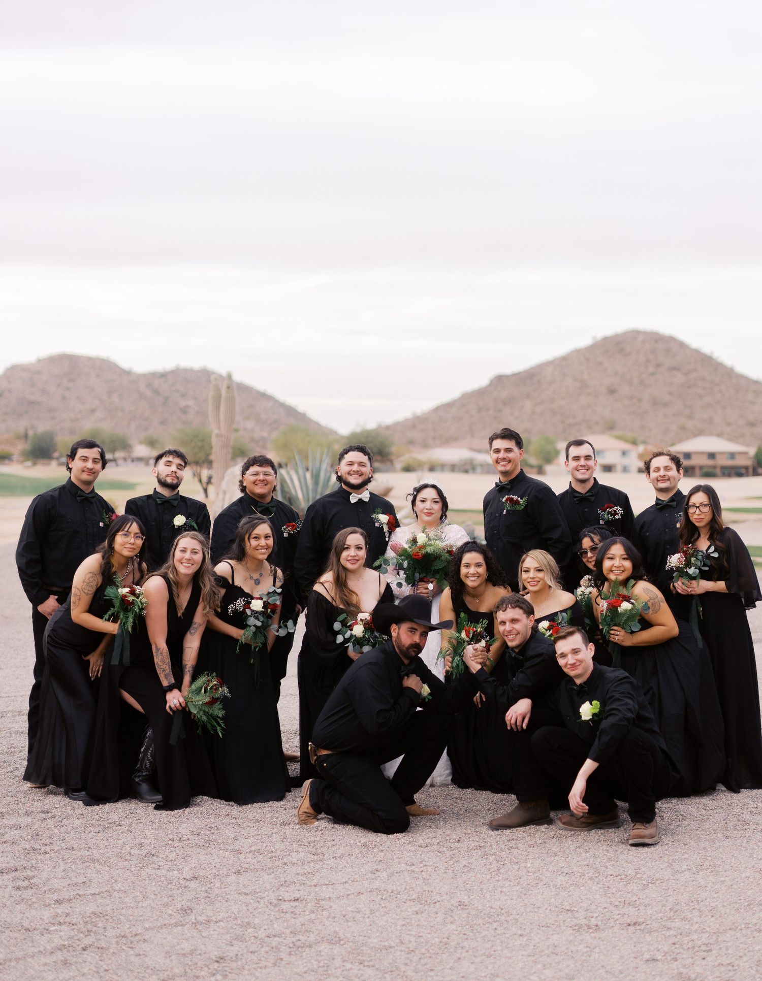Wedding party in all black attire poses together in desert setting with mountains in background at sunset.