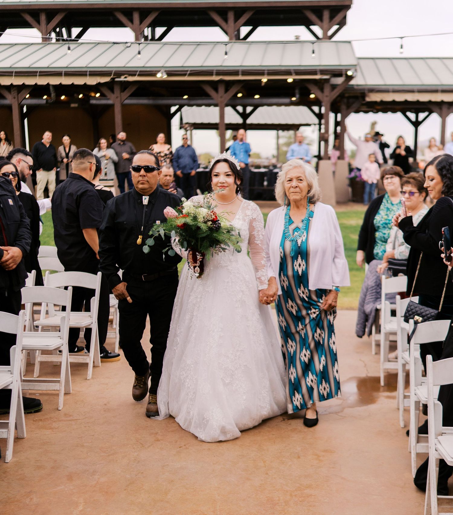 Bride in white wedding gown walks down outdoor aisle accompanied by guests at covered venue with white chairs.