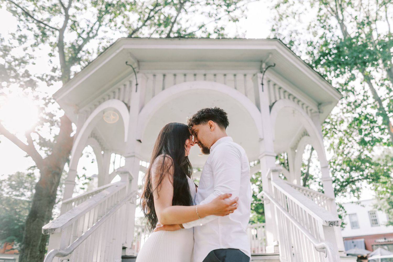 Couple sharing intimate moment under white gazebo with sunlight streaming through trees.