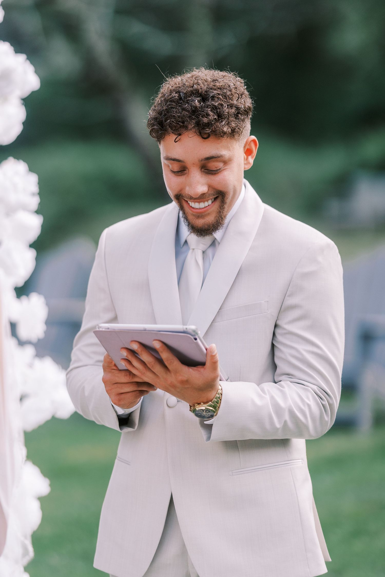 Groom in light gray suit reading from tablet during outdoor ceremony.