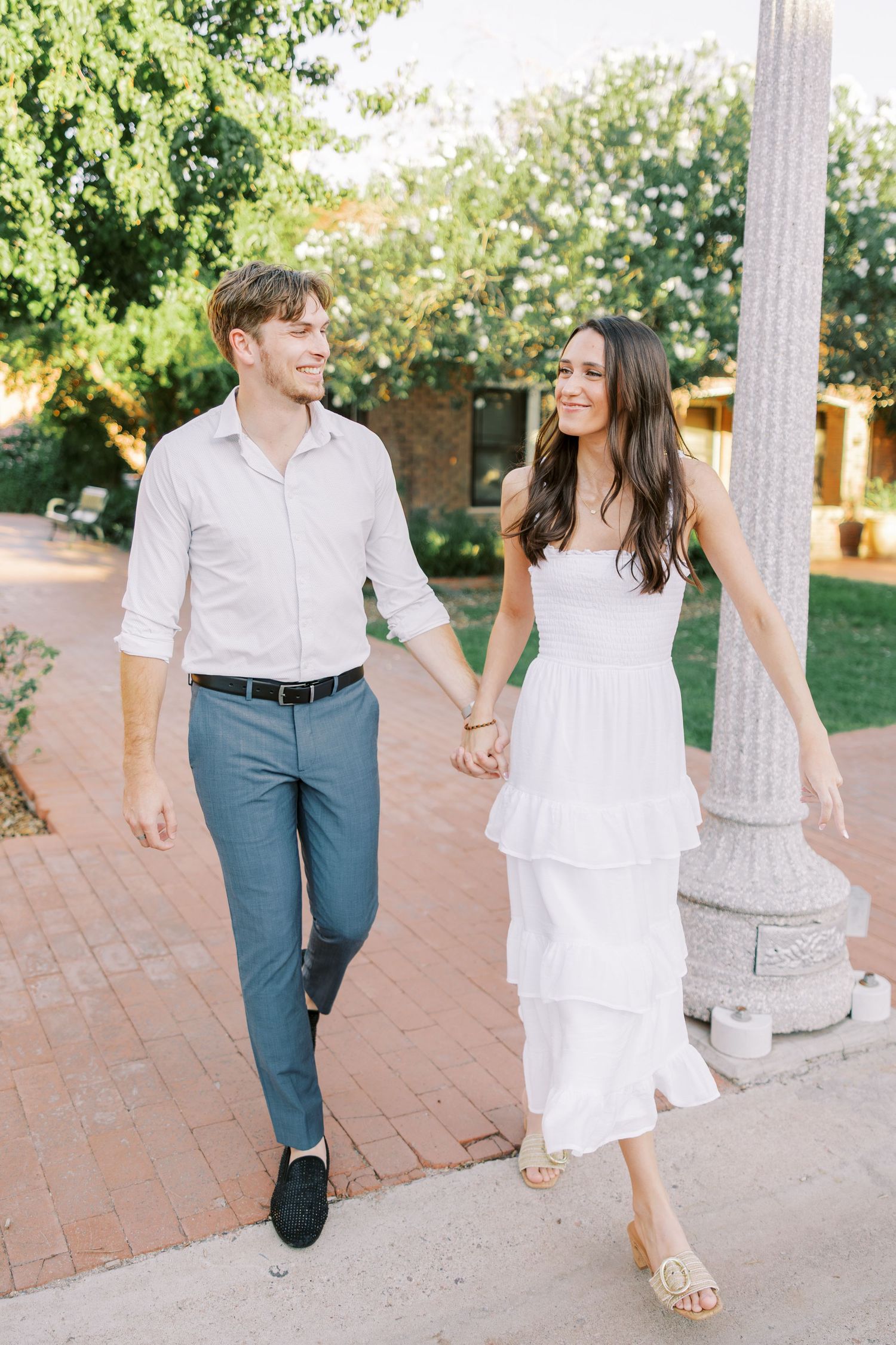 A couple walking together on a brick path with the woman wearing a flowing white dress and man in blue pants.