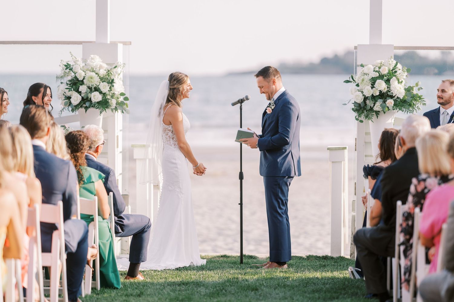 A scenic waterfront wedding ceremony takes place on a grassy lawn with white floral arrangements and seated guests.